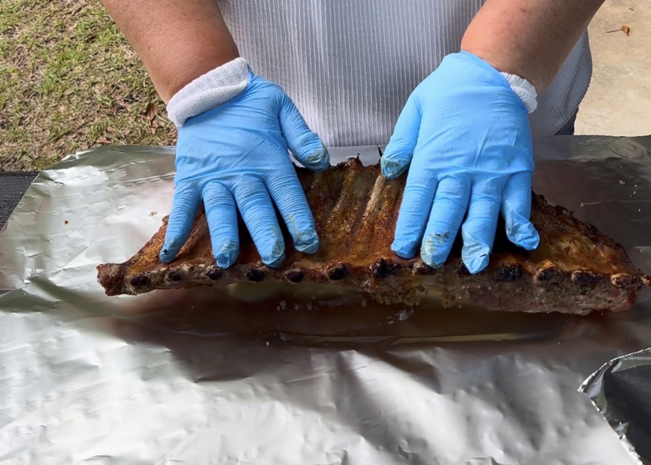 A person wearing blue gloves and a white shirt is handling a cooked rack of ribs on a sheet of aluminum foil outdoors.