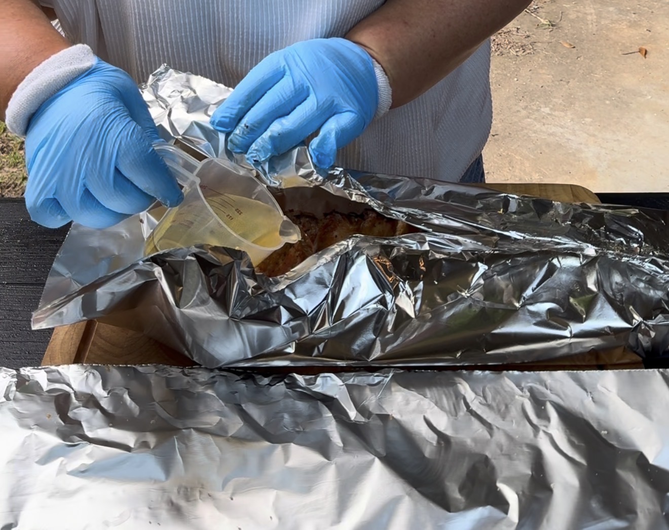 A person wearing blue gloves pours liquid from a small cup into a foil-wrapped dish on a wooden board outdoors, preparing food.