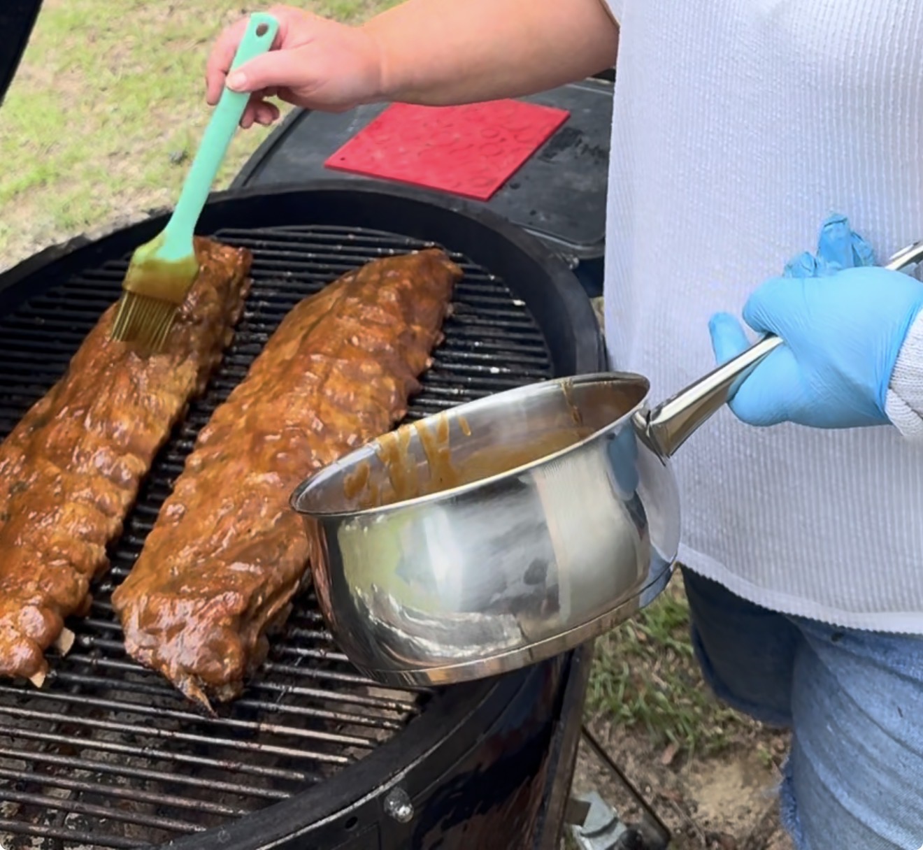 A person wearing a white shirt and blue gloves brushes sauce onto two racks of ribs cooking on an outdoor grill, holding a pot of sauce in the other hand.