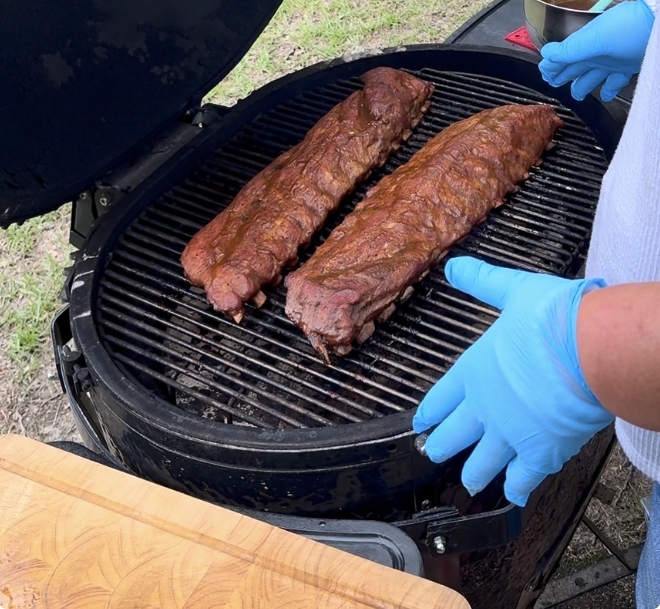 Two racks of seasoned ribs cooking on a round charcoal grill outdoors. A person wearing blue gloves is tending the grill, while a wooden cutting board sits nearby.