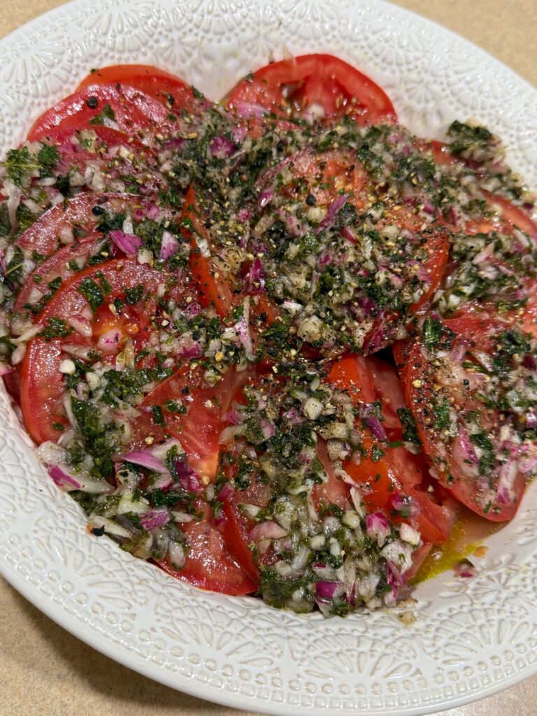 A white plate filled with sliced tomatoes topped with chopped red onions, herbs, and cracked black pepper, dressed with oil and seasoning.