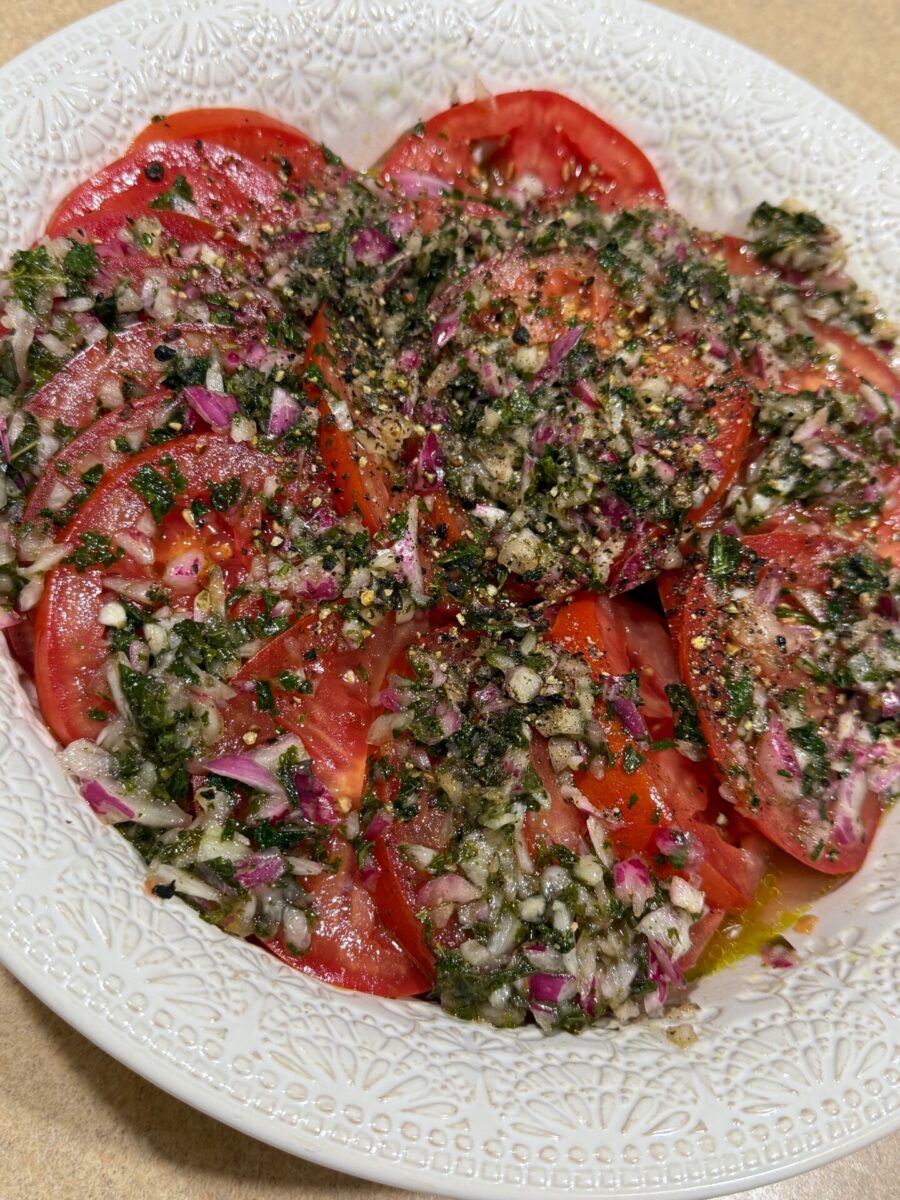 A white plate filled with sliced tomatoes topped with chopped red onions, herbs, and cracked black pepper, dressed with oil and seasoning.