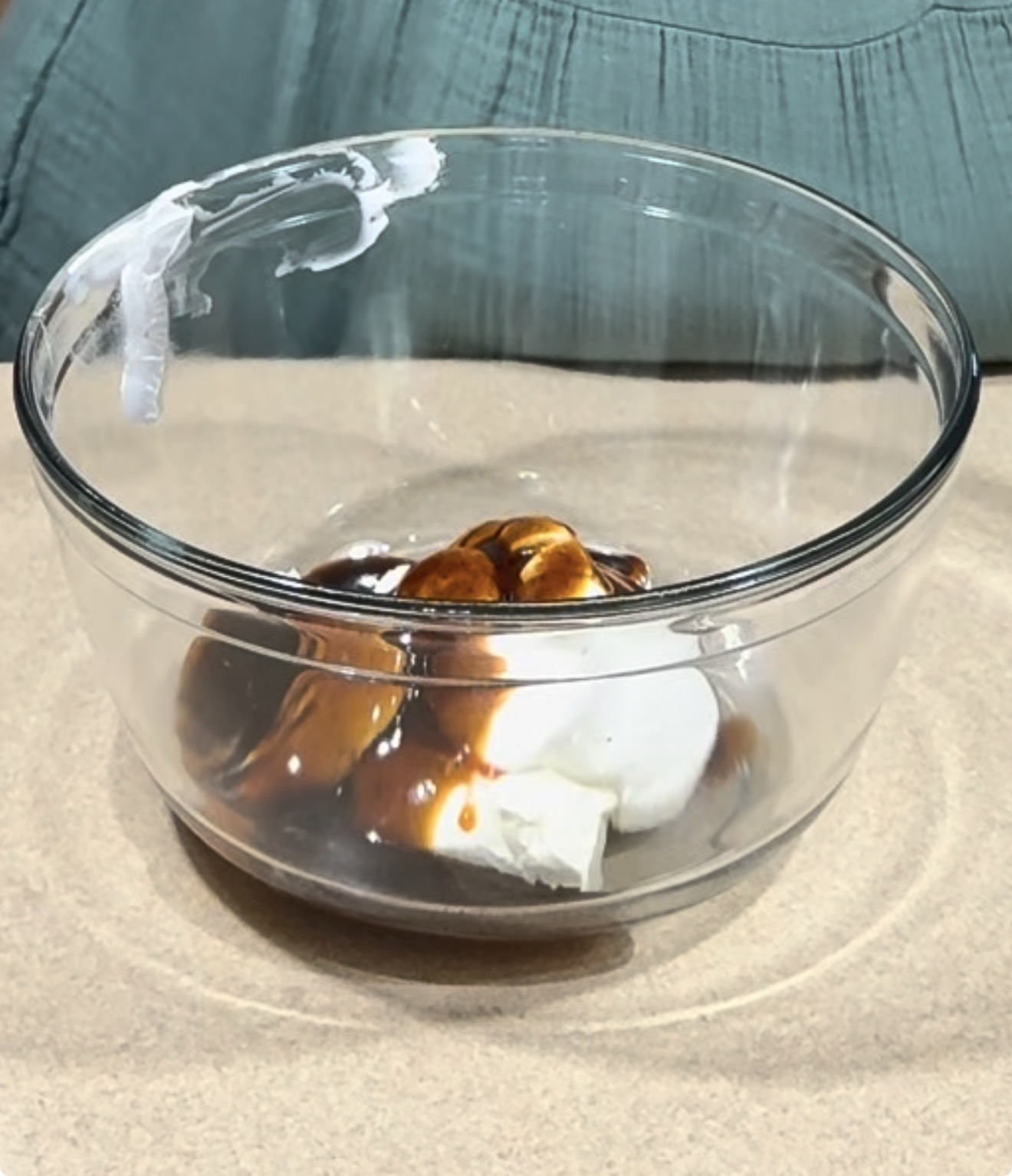 A clear glass bowl containing white sour cream and brown sauce sits on a beige surface with a person in a green shirt in the background.