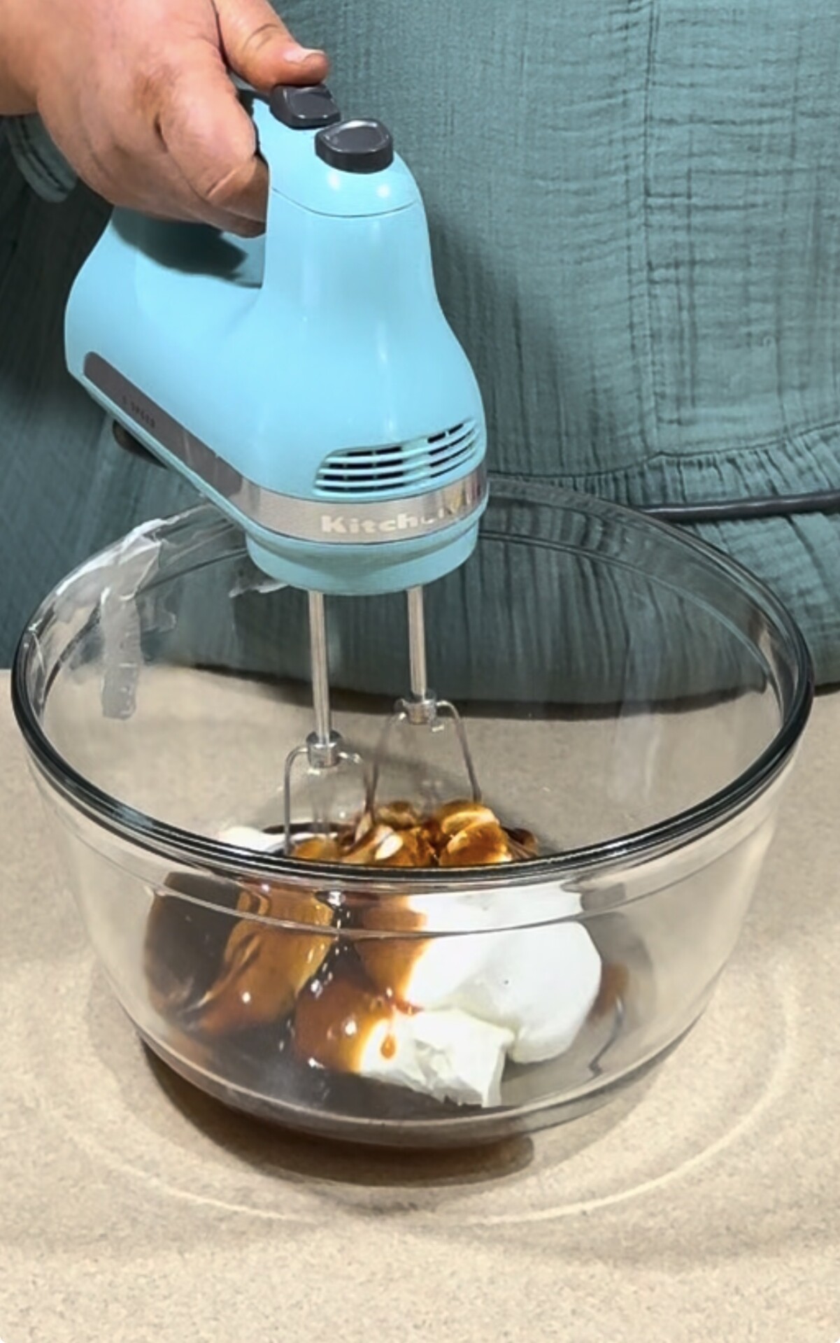 A person uses a light blue hand mixer to blend ingredients, including sour cream and vanilla, in a clear glass mixing bowl on a kitchen counter.