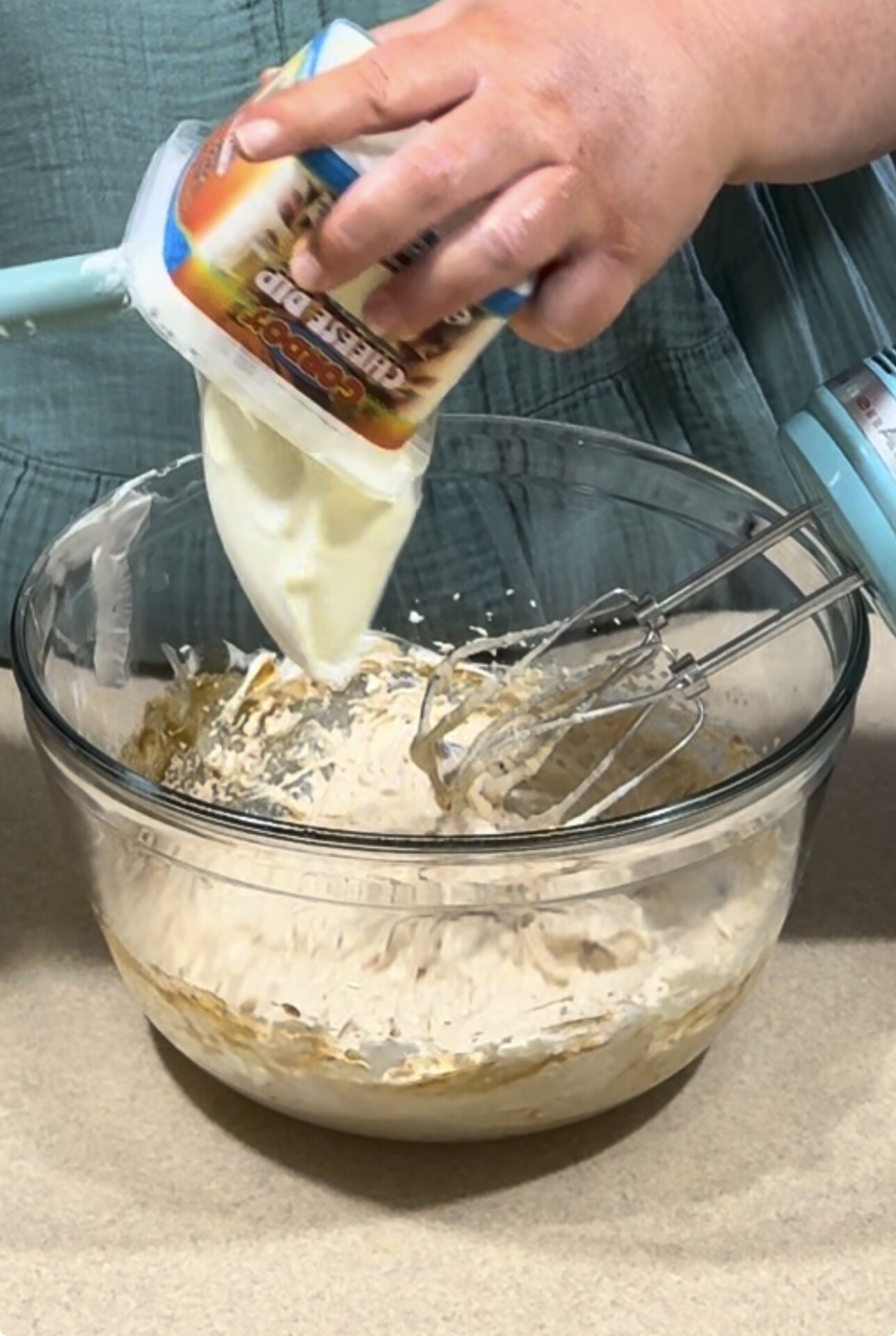 A person pours cheesecake dip from a container into a glass mixing bowl with a hand mixer and batter inside, preparing ingredients on a beige countertop.
