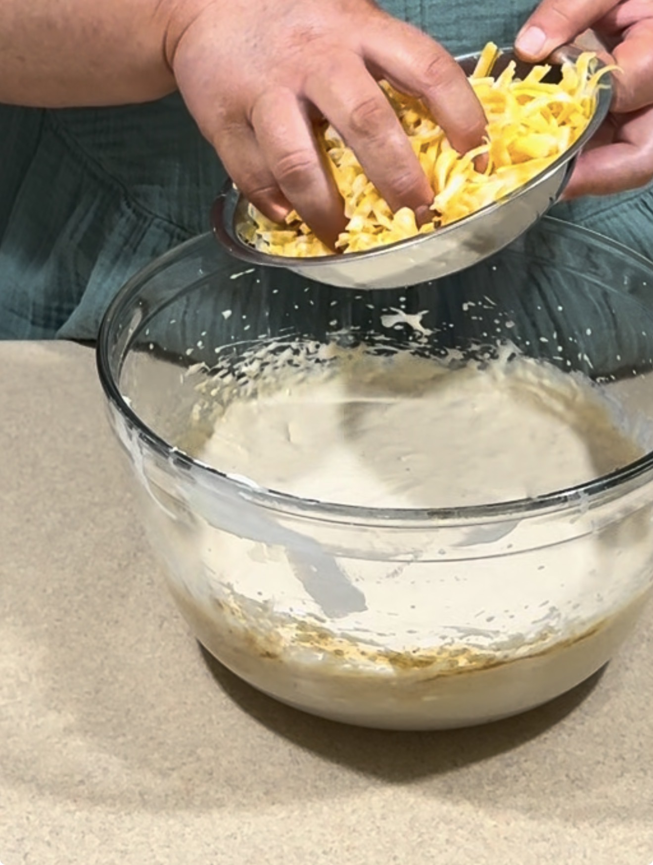 A person adds shredded cheese from a small bowl into a larger glass bowl containing a creamy batter mixture on a countertop.