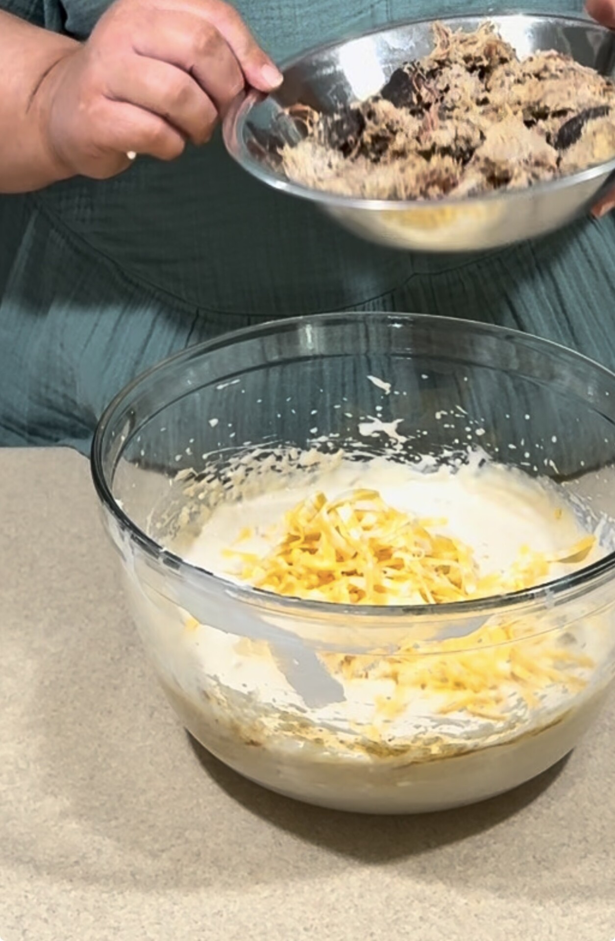 A person in a teal shirt holds a metal bowl with shredded meat above a glass bowl containing a creamy mixture and shredded cheese on a countertop.
