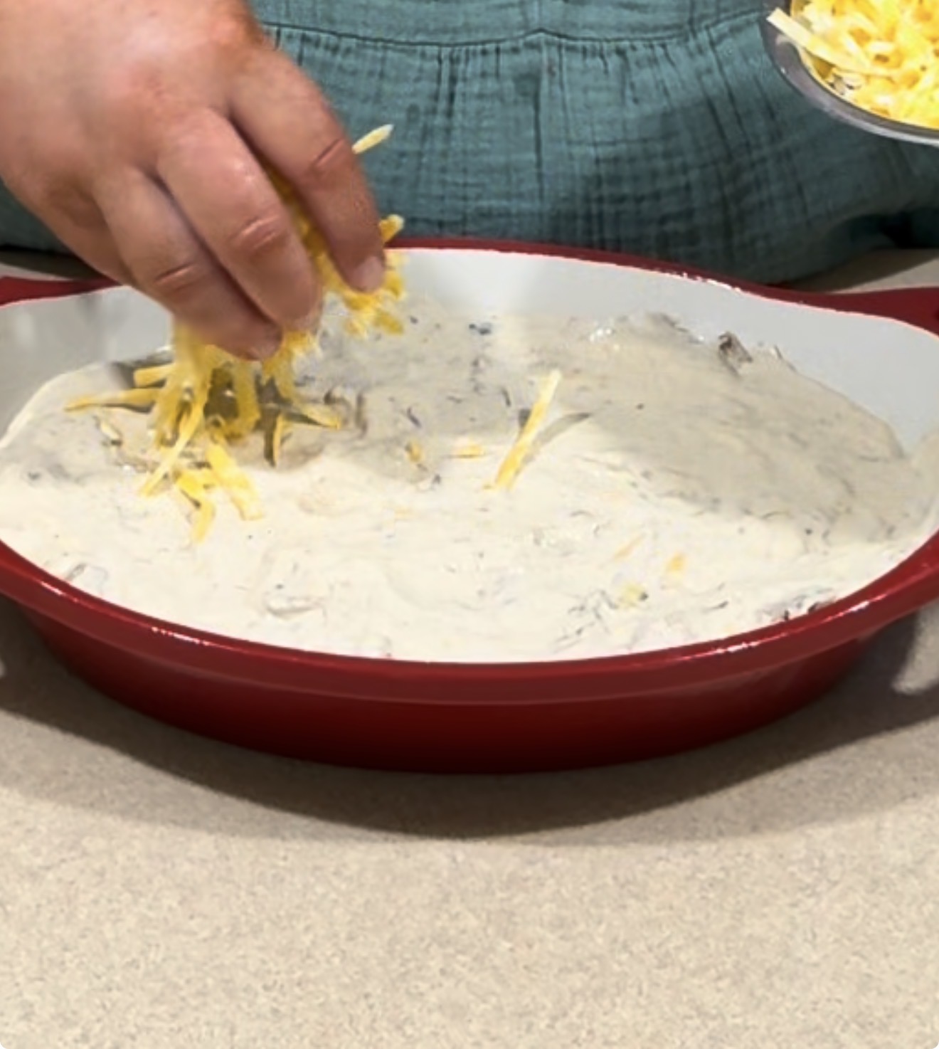 A person sprinkles shredded cheese over a creamy mixture in a red oval baking dish on a countertop.