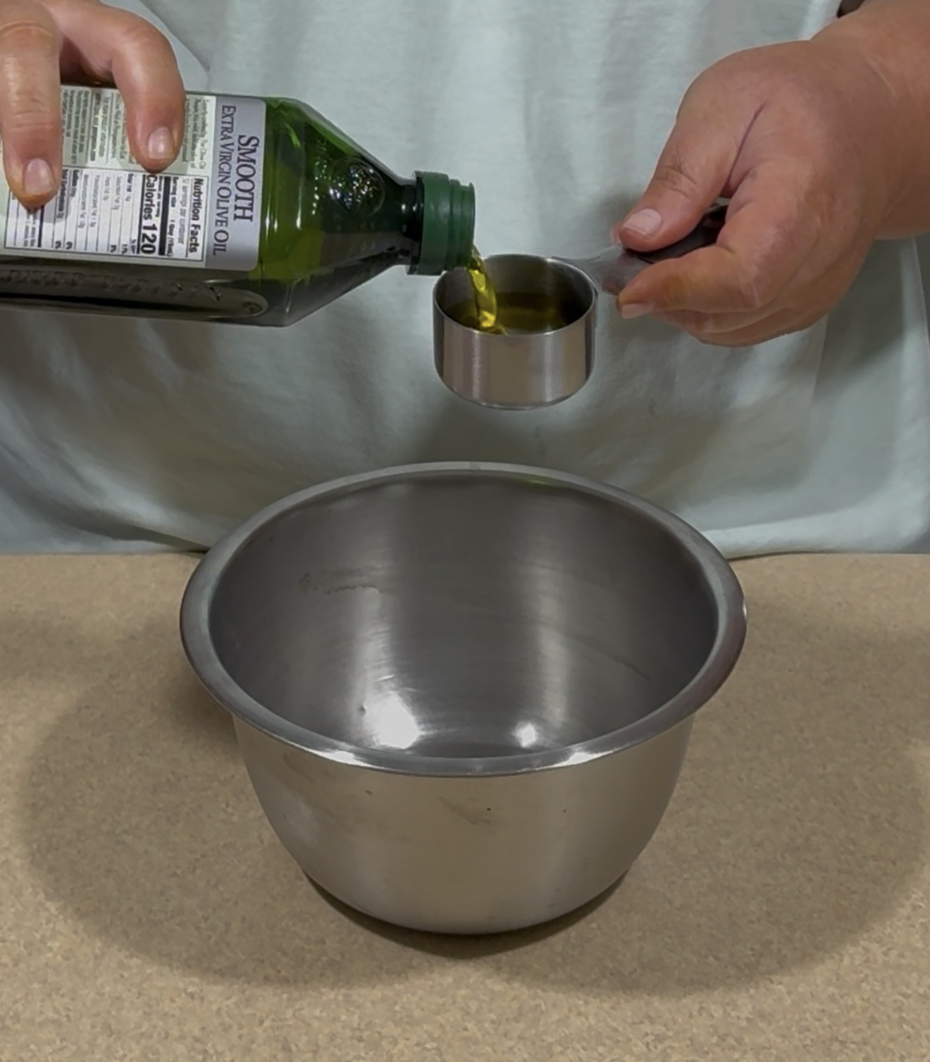 A person pours olive oil from a bottle into a metal measuring cup over a large empty metal mixing bowl on a kitchen counter.