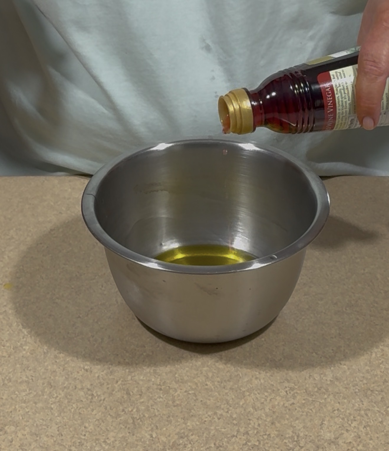 A person pours liquid from a small bottle into a stainless steel bowl containing yellow oil, set on a countertop.