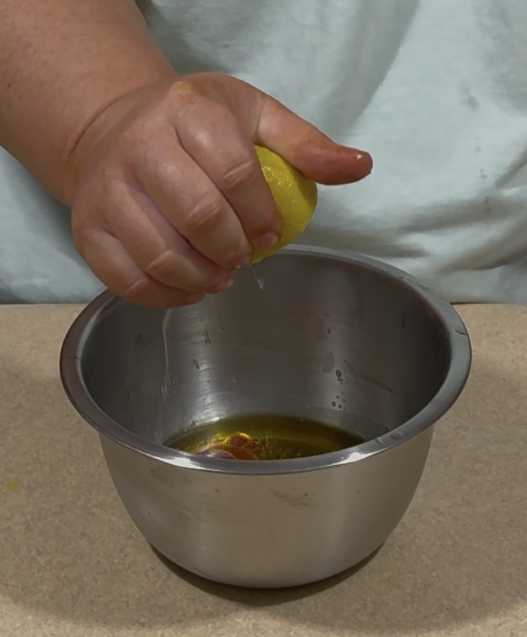 A person squeezing a lemon over a metal bowl containing liquid, likely preparing ingredients for a recipe on a countertop.
