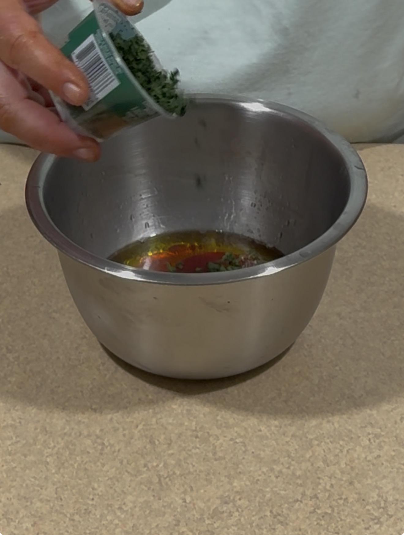 A person adds chopped herbs from a small container into a stainless steel mixing bowl with various liquid ingredients on a beige countertop.