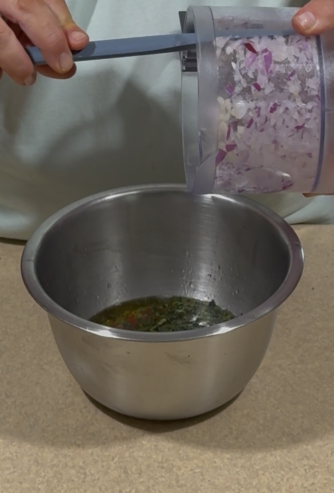 A person pours chopped red onions from a food processor into a metal mixing bowl containing a green and yellow liquid mixture on a countertop.
