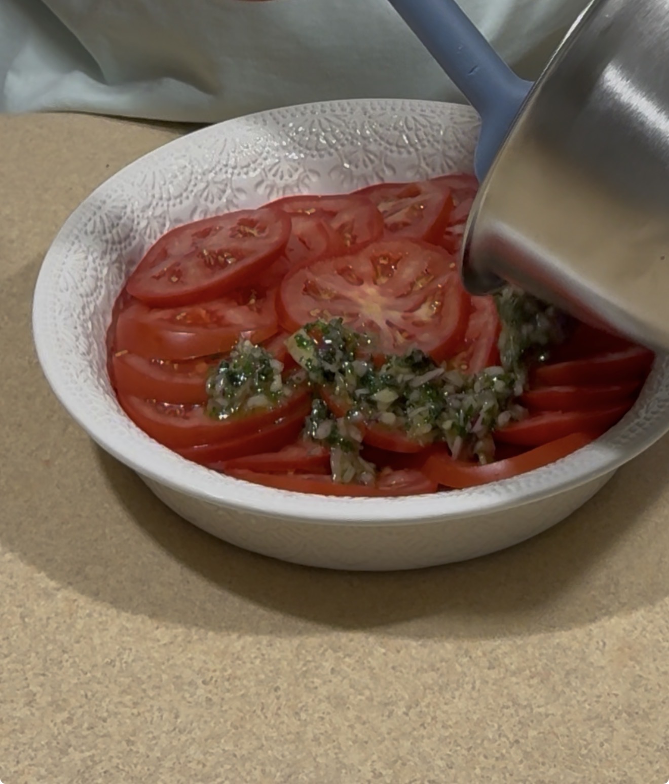A person pours a herb and oil mixture from a saucepan onto sliced tomatoes arranged in a white textured dish.