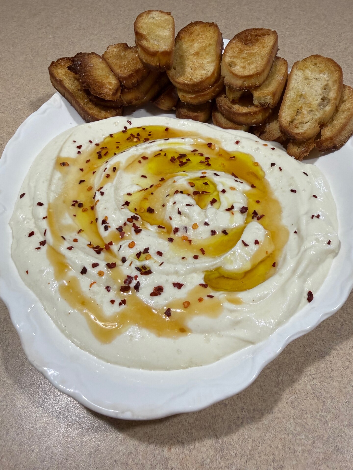 A plate of creamy white dip topped with olive oil and red pepper flakes, served with a stack of toasted bread slices on the side.