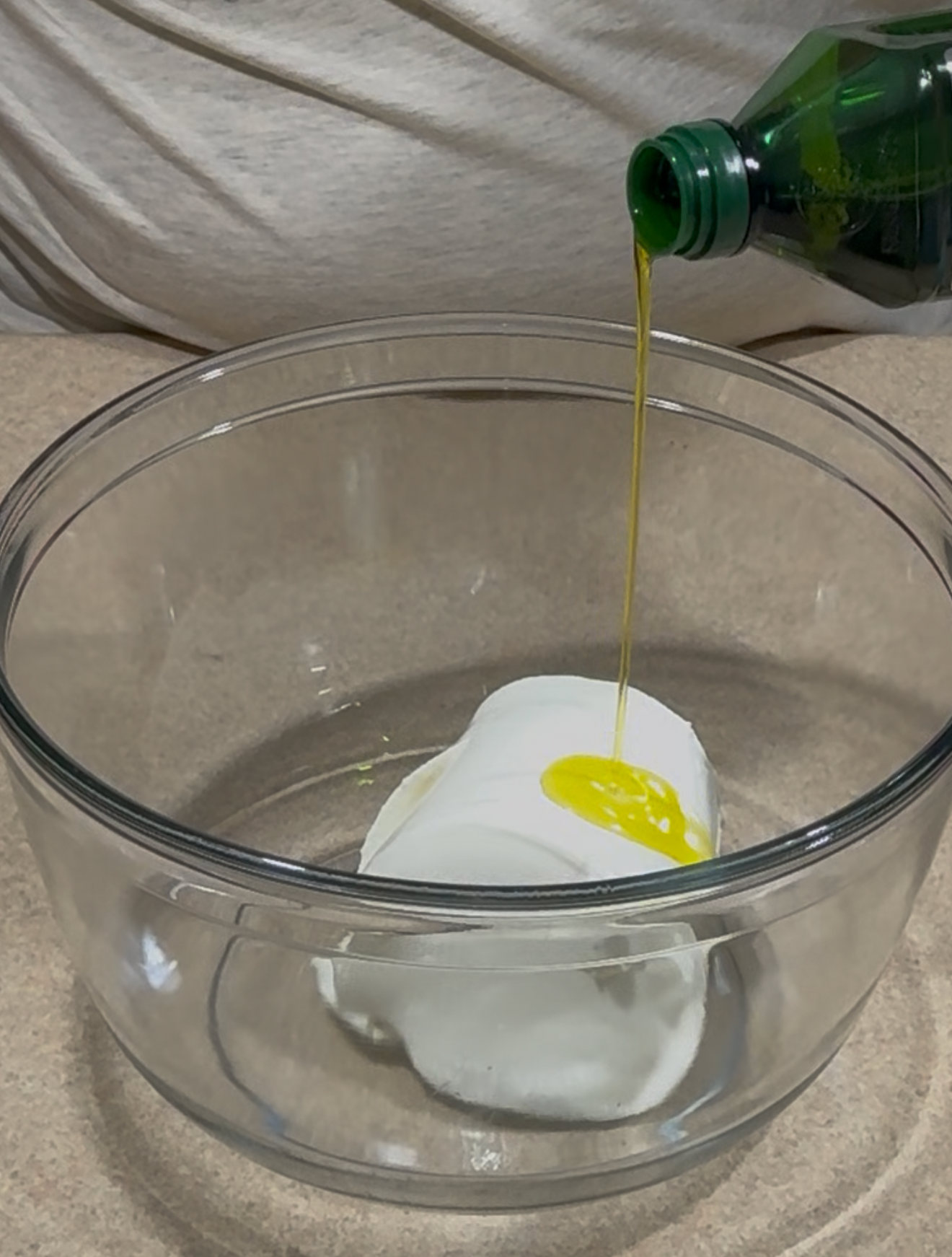 A person pours olive oil from a green bottle into a glass bowl containing a large scoop of white solid, possibly shortening or lard, on a kitchen counter.