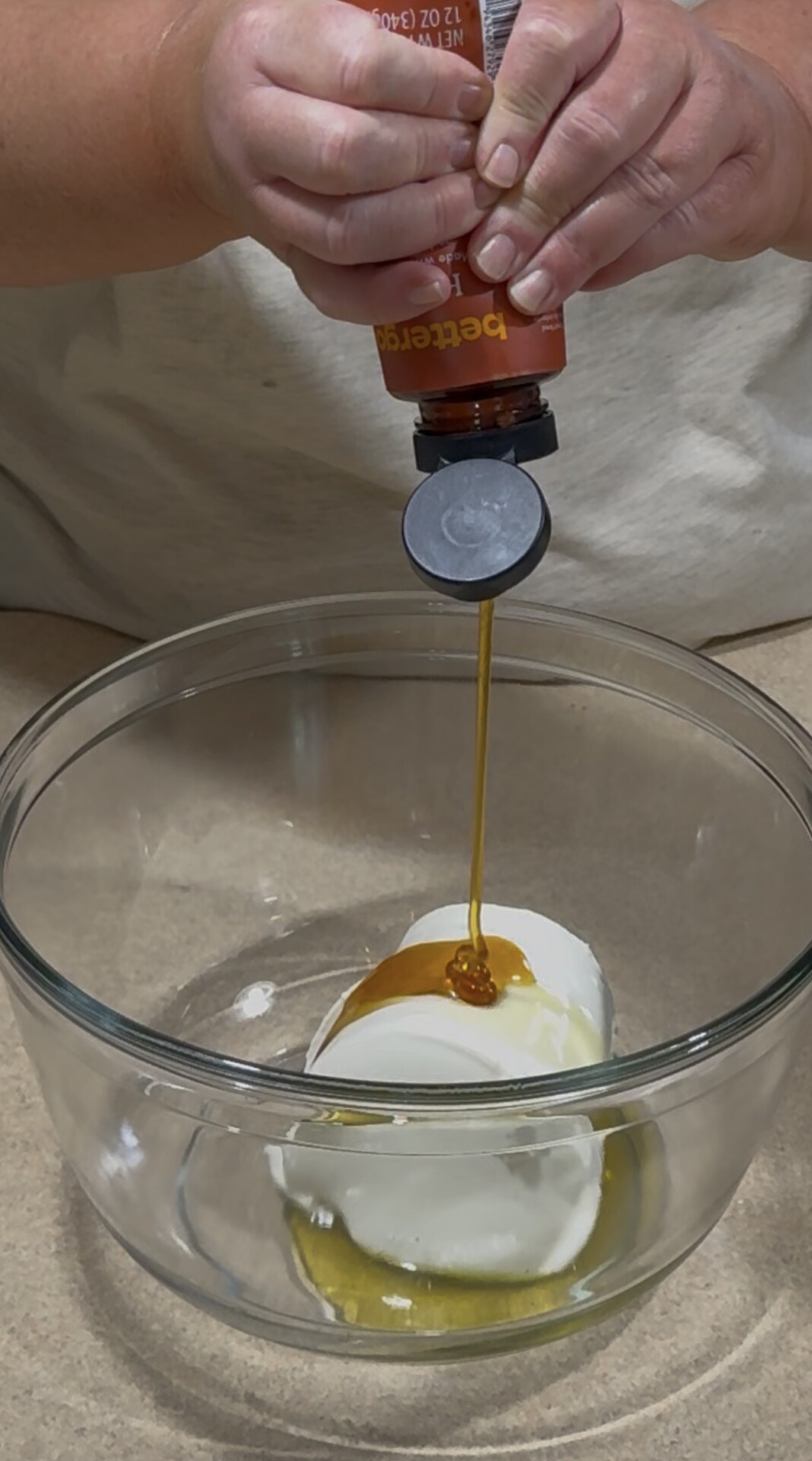 A person squeezes honey from a bottle onto a block of cream cheese in a glass mixing bowl on a kitchen counter.