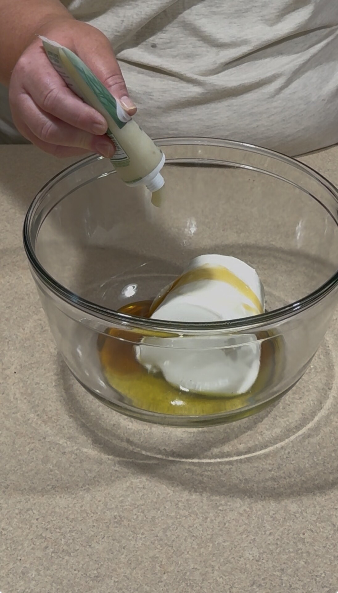 A person squeezes liquid from a tube into a glass bowl containing white yogurt or sour cream and golden syrup or honey on a beige countertop.