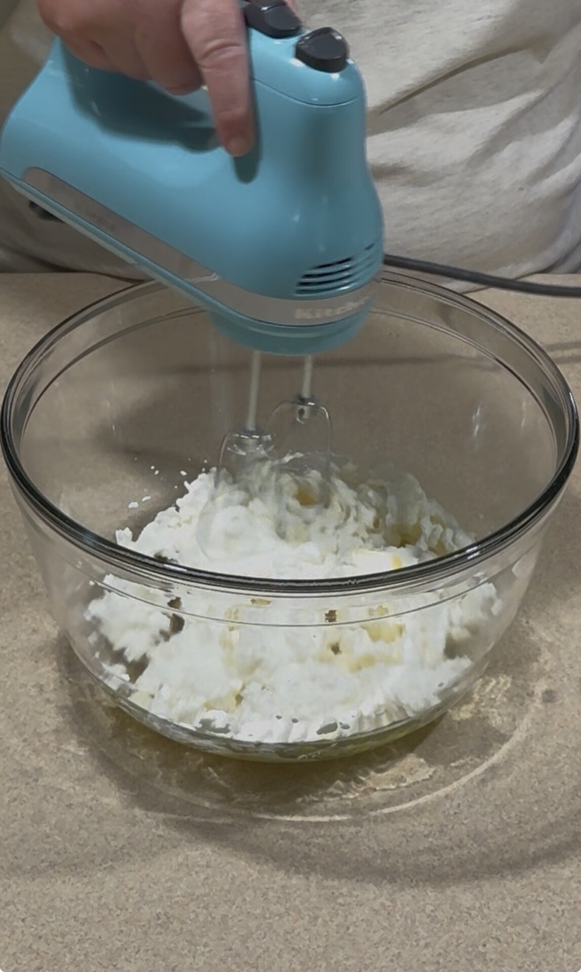 A person uses a blue electric hand mixer to blend ingredients, including whipped cream and liquid, in a large glass bowl on a kitchen countertop.