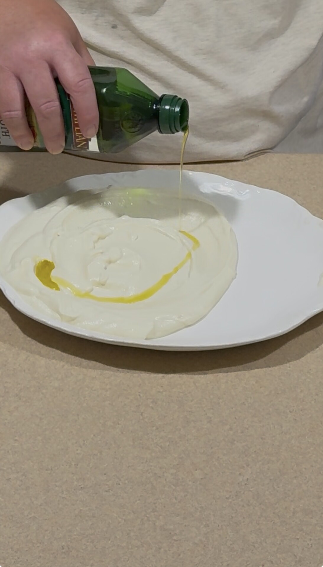 A hand pours olive oil from a bottle onto a plate of creamy white yogurt spread, with both placed on a beige countertop.
