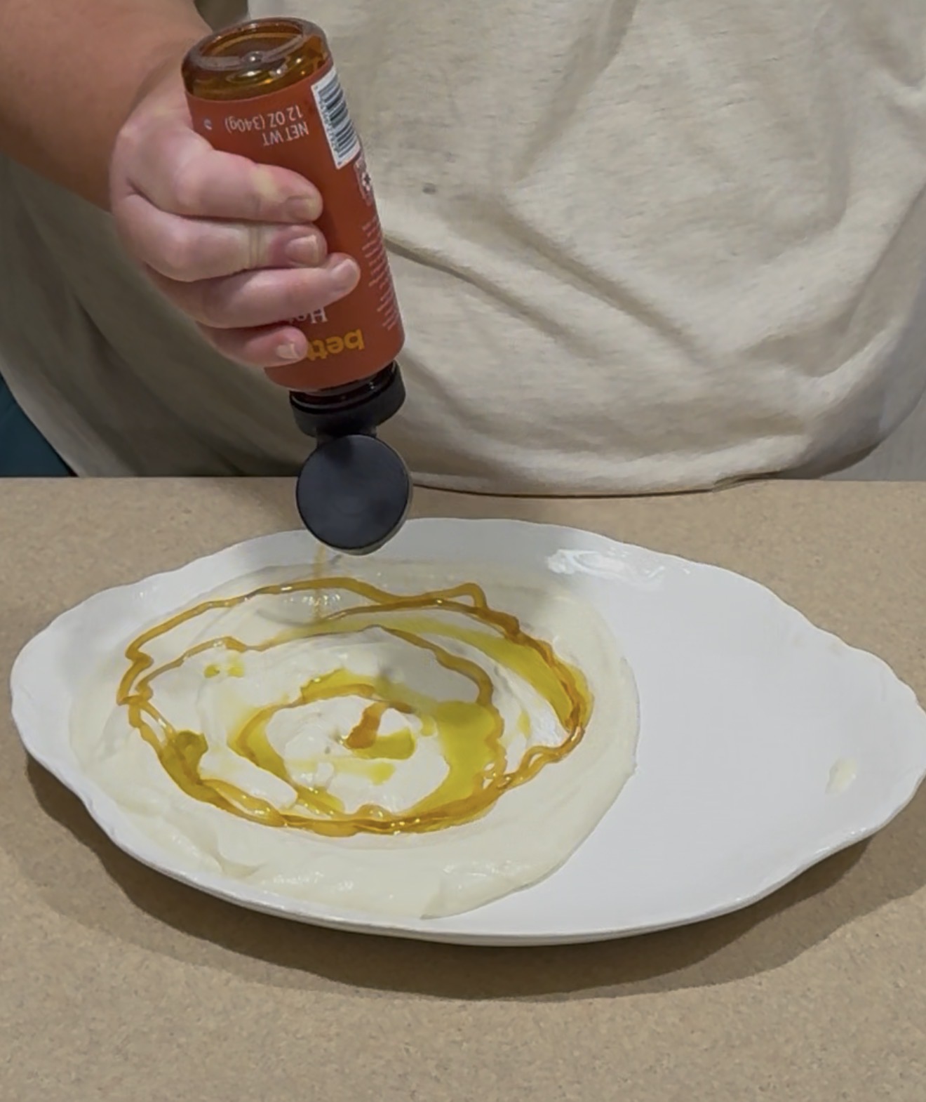 A person pours honey from a bottle onto a plate of creamy white yogurt, creating a swirl pattern on the surface. The plate is set on a beige countertop.