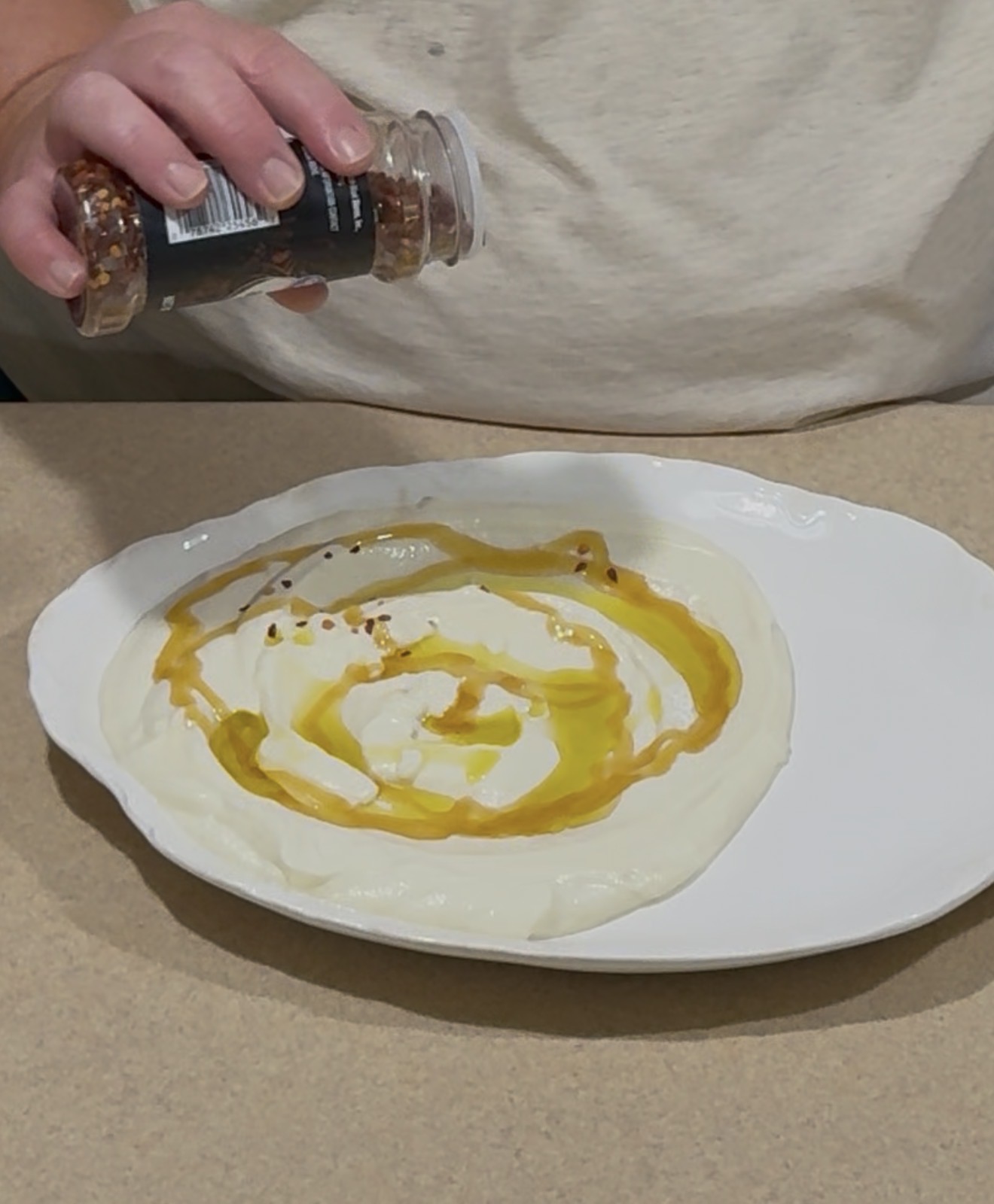 A person sprinkles red pepper flakes from a jar onto a plate of creamy yogurt drizzled with olive oil.