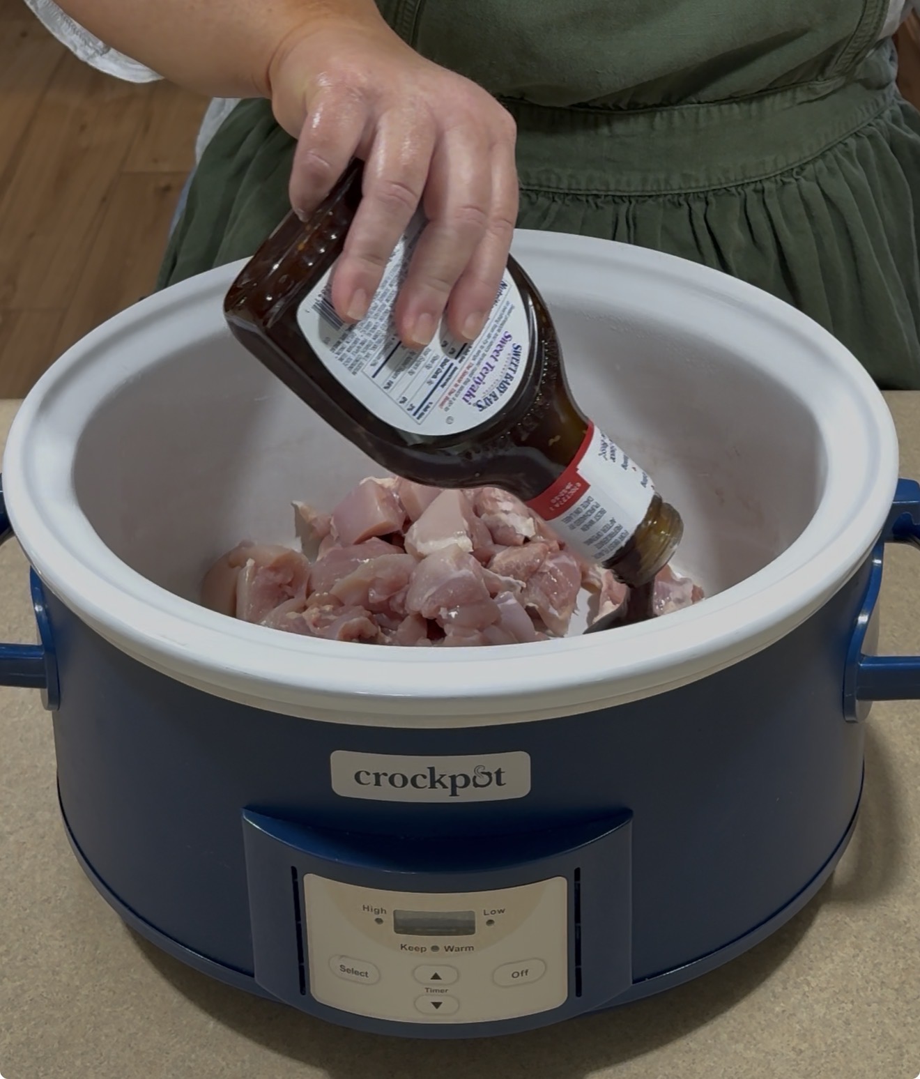 A person pours barbecue sauce from a bottle onto raw, chopped chicken pieces inside a blue Crock-Pot slow cooker on a kitchen counter.