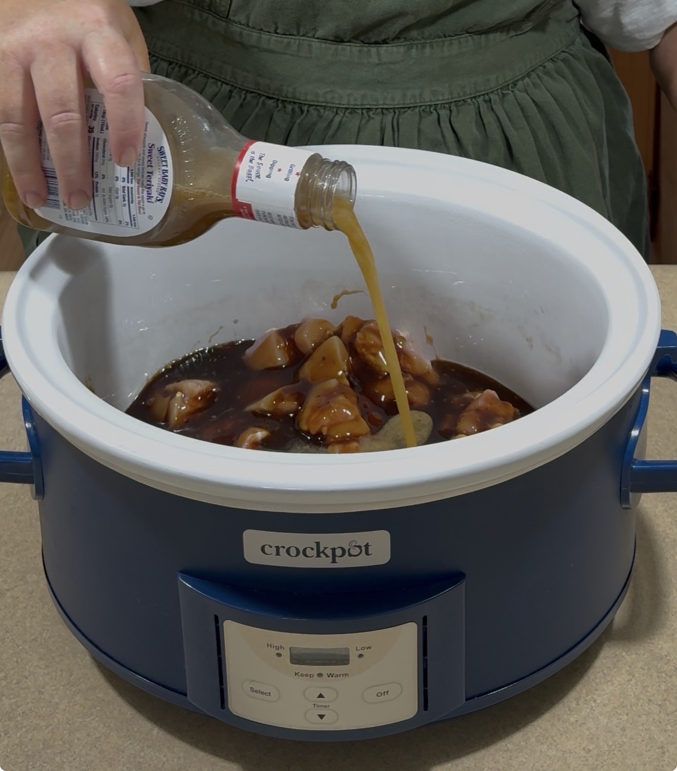 A person pours sauce from a bottle onto pieces of raw chicken in a blue Crock-Pot slow cooker placed on a kitchen counter.