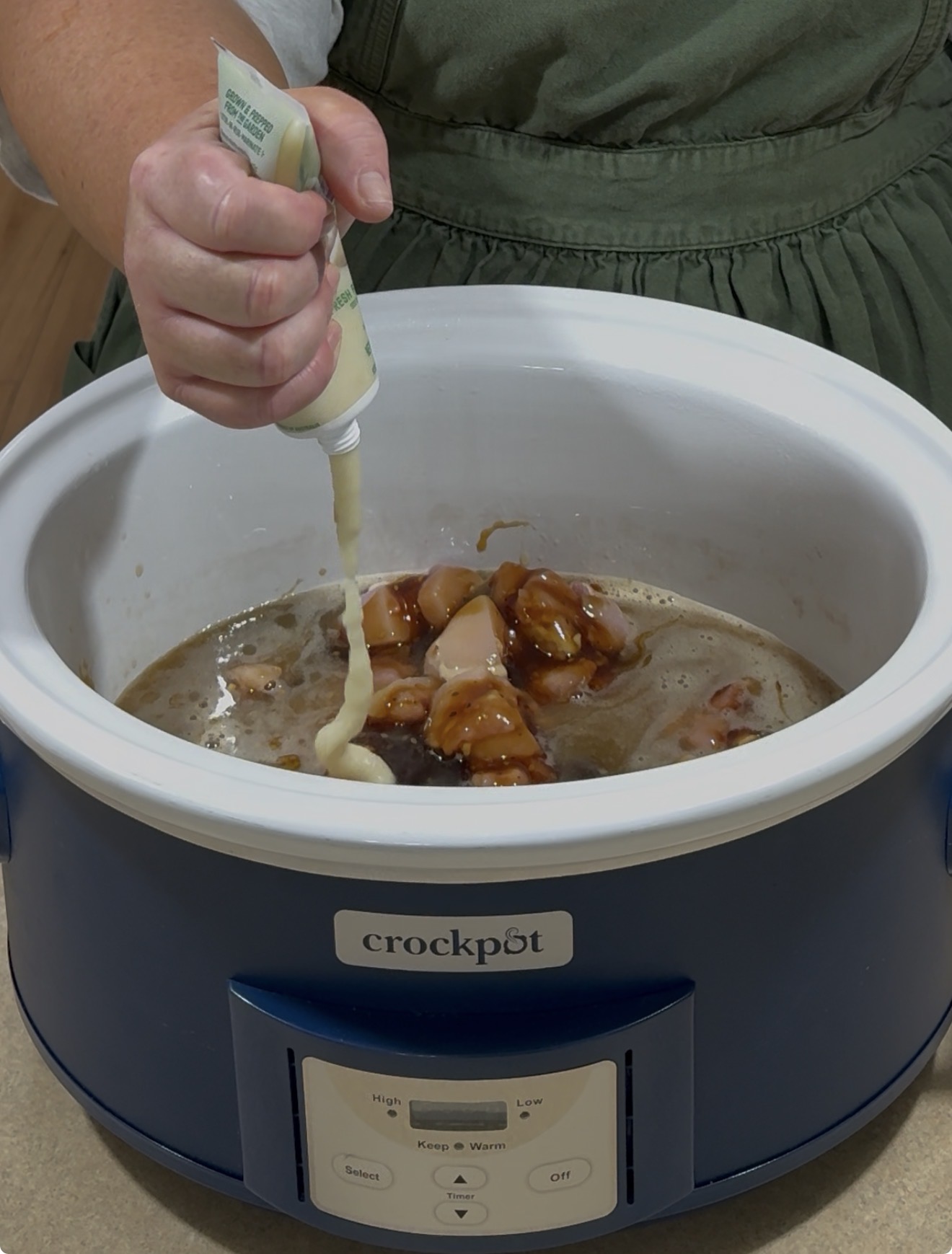 A person squeezes a tube of seasoning or sauce into a blue Crockpot filled with chicken pieces and sauce on a kitchen counter.
