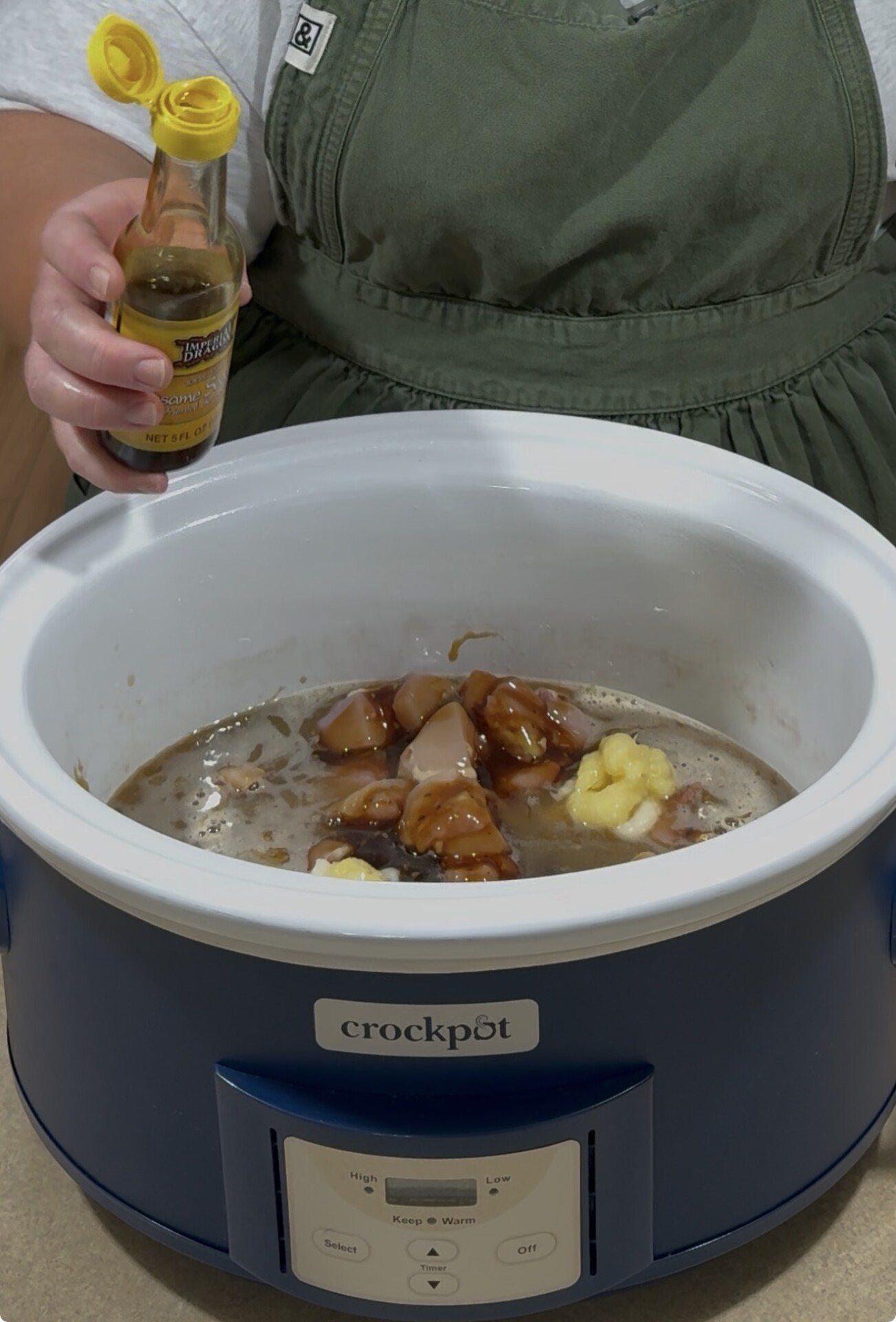 A person in a green apron pours Worcestershire sauce from a bottle into a slow cooker filled with chunks of meat, sauce, and butter. The slow cooker is blue and labeled “crockpot.”.