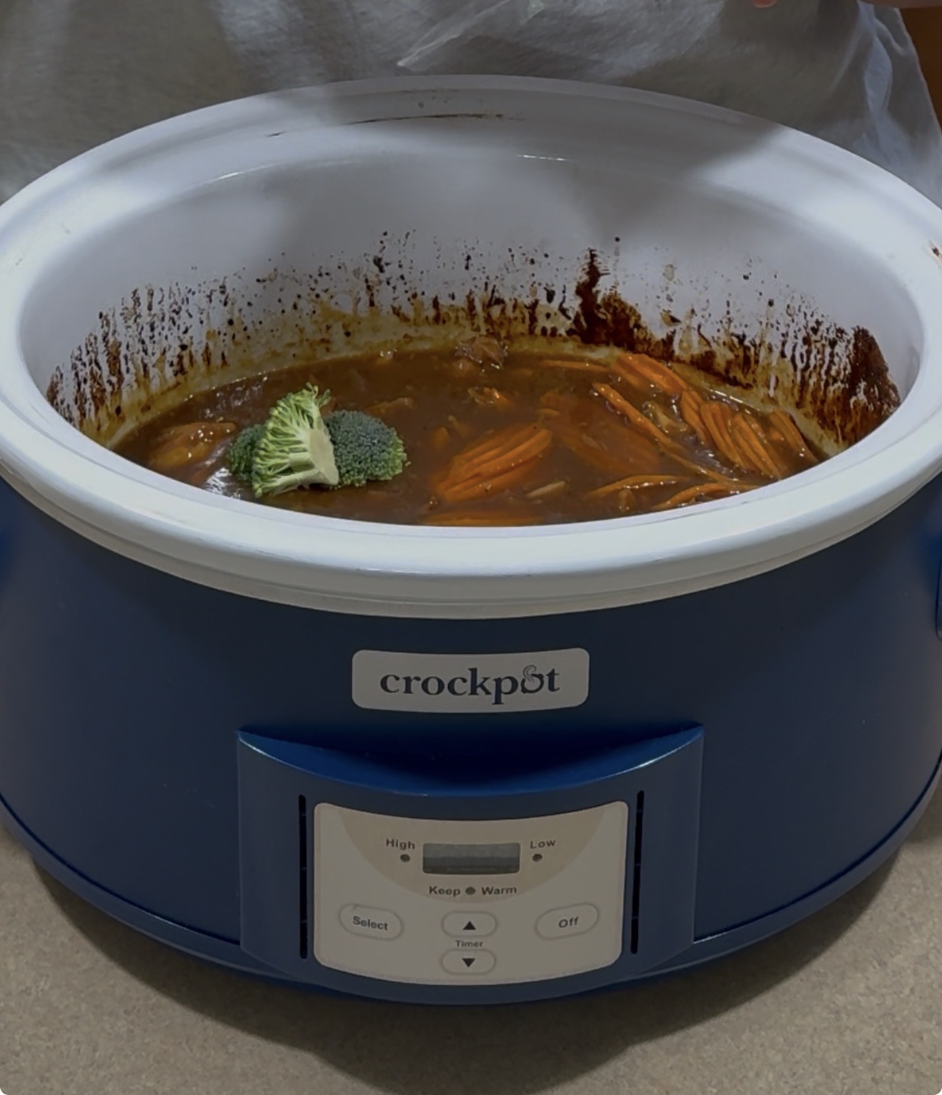 A blue Crock-Pot slow cooker containing a stew with visible shredded meat, sauce, carrots, and a piece of broccoli on top, sitting on a kitchen counter.