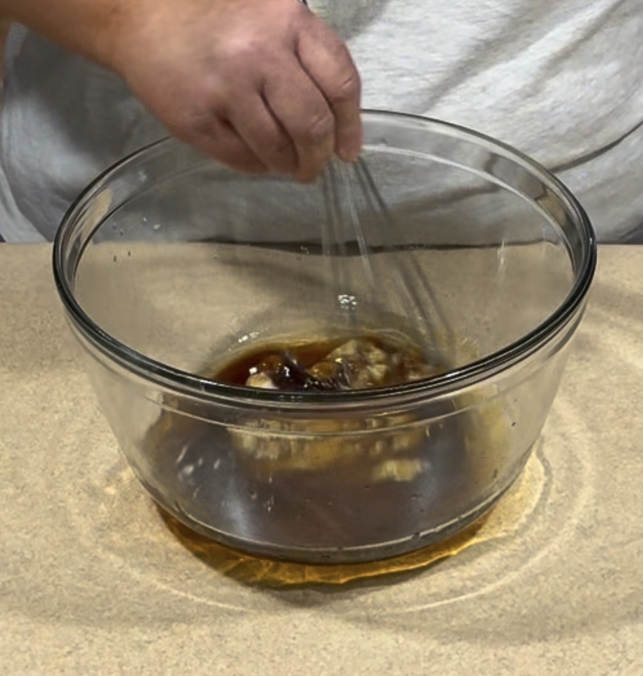 A person is whisking a dark liquid mixture in a large clear glass bowl on a beige countertop.