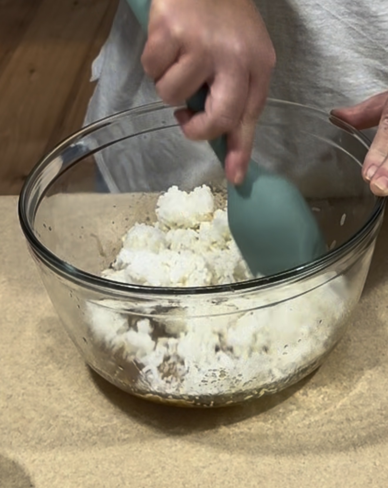 A person uses a teal spatula to mix white rice in a clear glass bowl on a countertop.