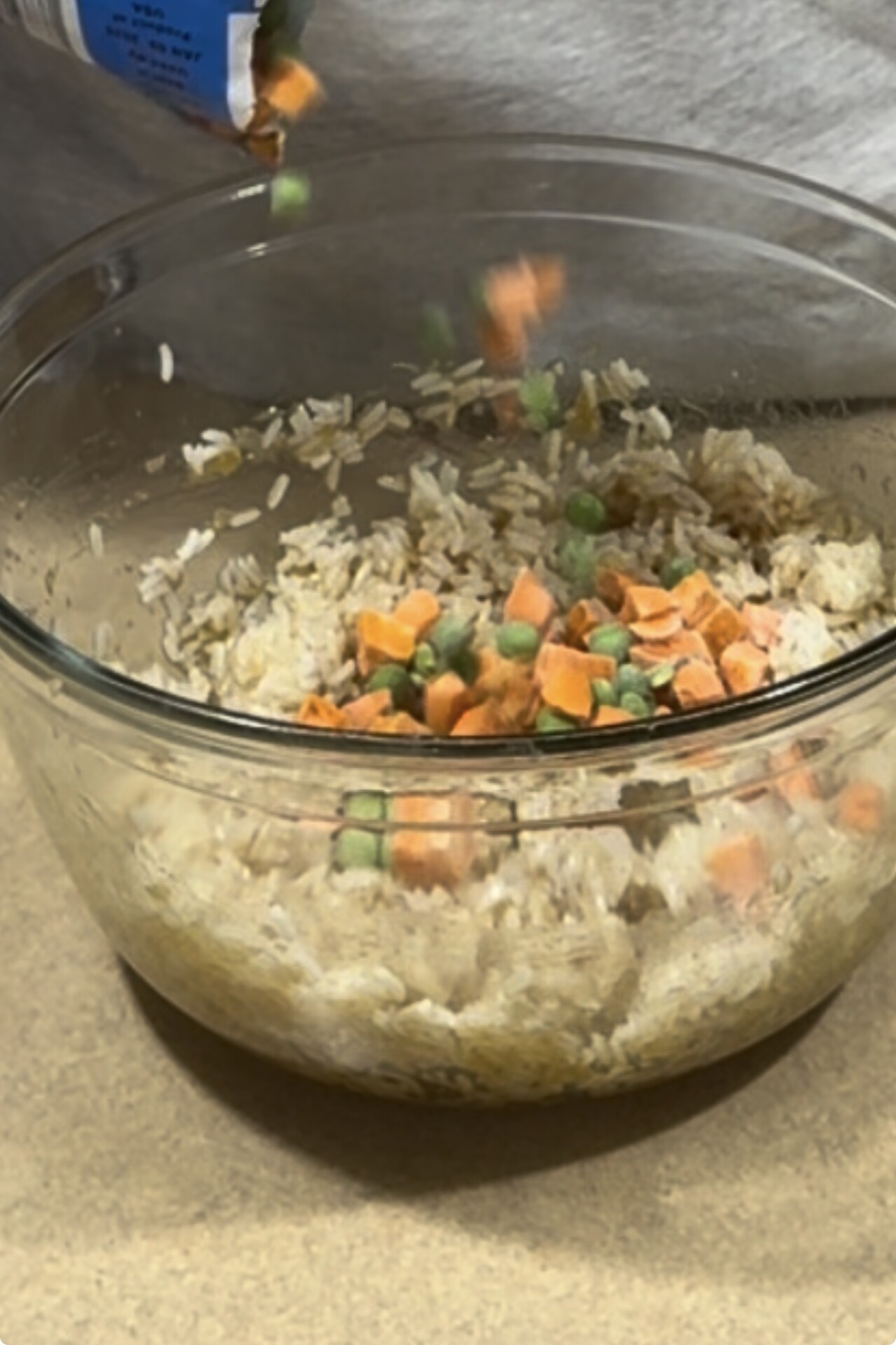 A glass bowl filled with rice sits on a counter as a person pours a mixture of frozen peas and diced carrots into it.