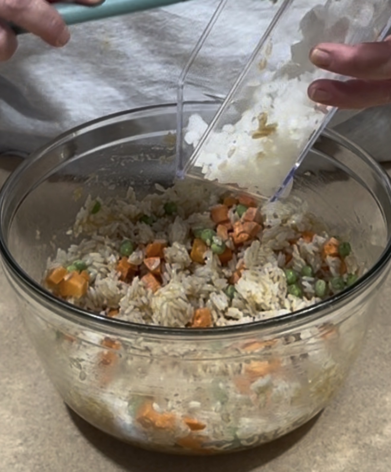 A person adds white rice from a container into a glass bowl filled with rice, peas, and diced carrots.
