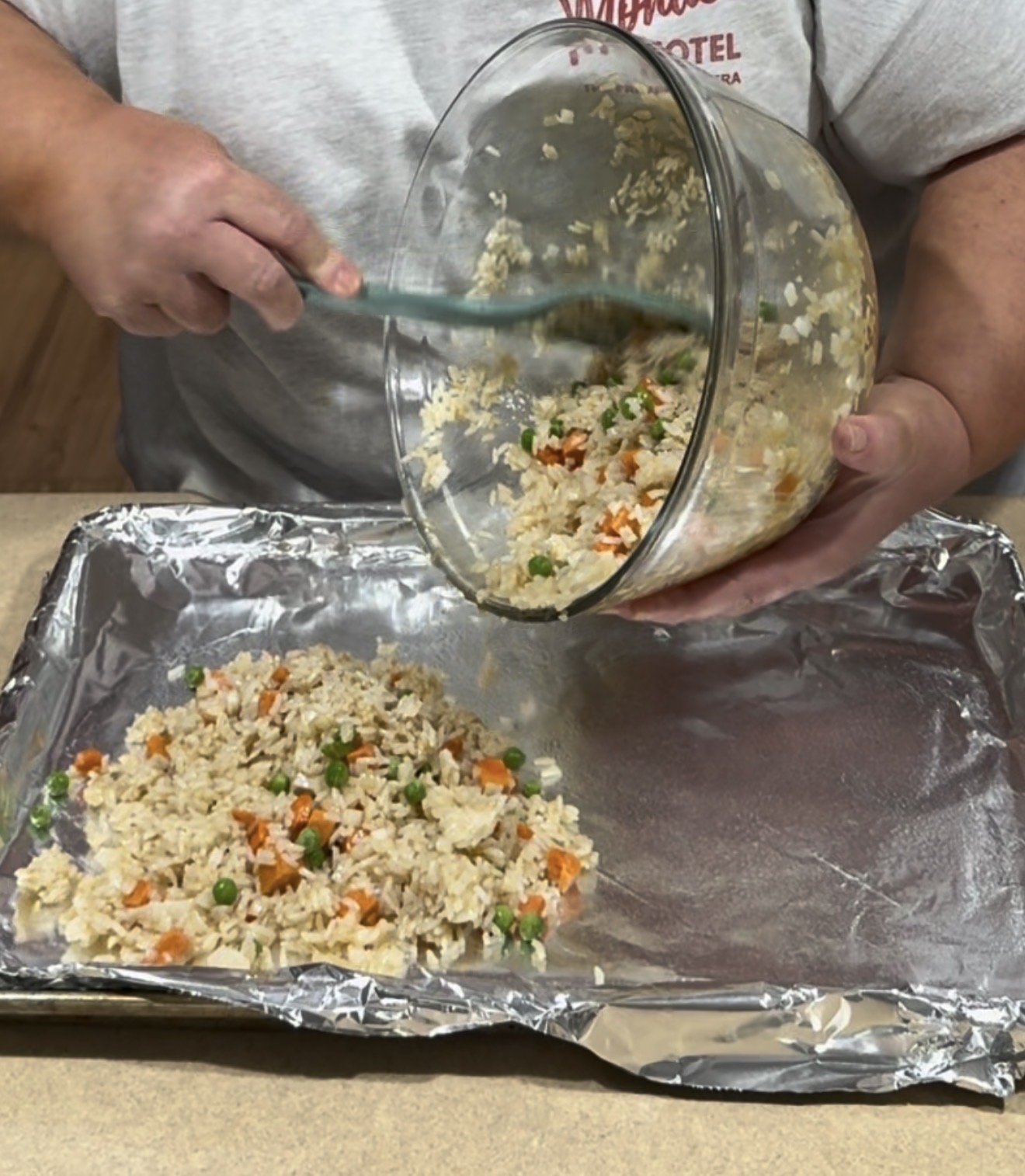 A person pours rice mixed with peas and carrots from a glass bowl onto a foil-lined baking sheet using a spatula.
