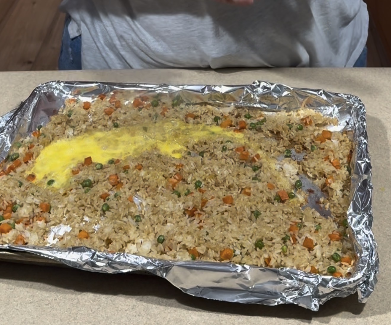 A foil-lined baking sheet on a countertop holds fried rice with peas and carrots, and a raw beaten egg spread over part of the rice. A person stands behind the sheet, partly visible from the shoulders down.