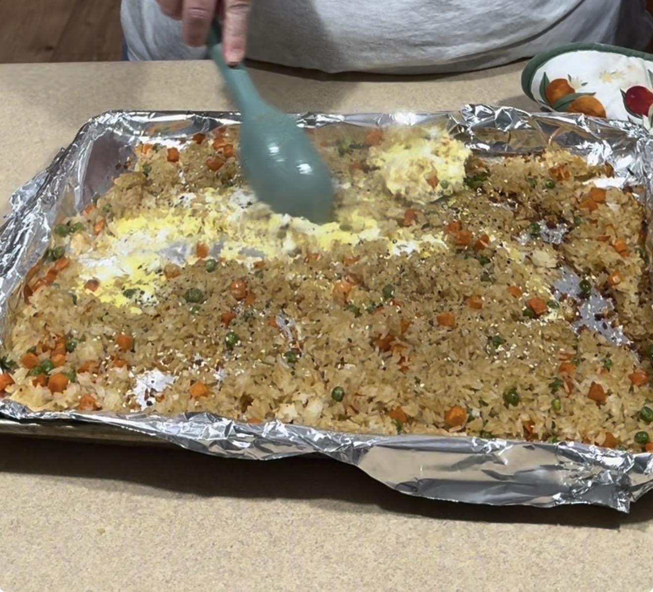 A person stirs vegetables and fried rice with a blue spatula on a foil-lined baking sheet. The rice contains peas and diced carrots, and there is a bowl with cut vegetables nearby on the counter.