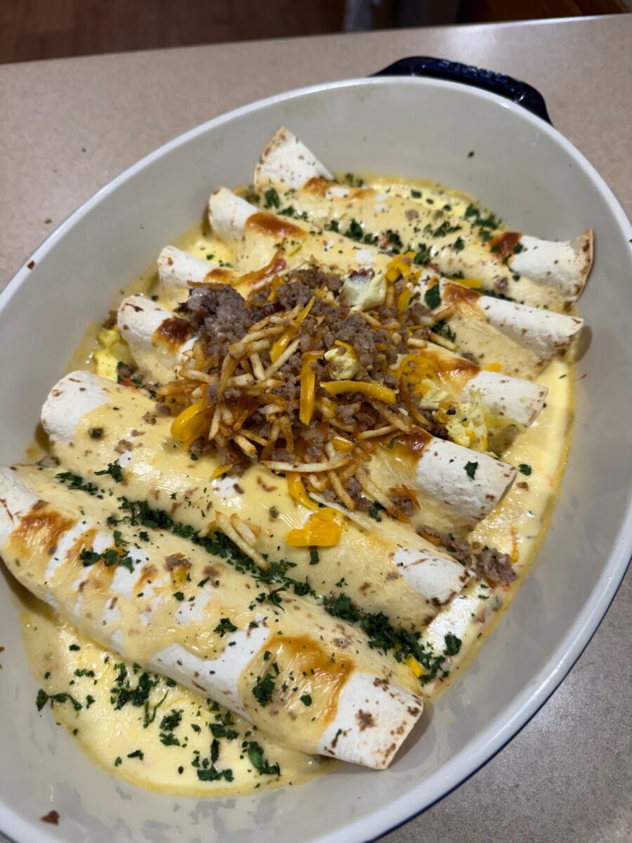 A white baking dish filled with baked rolled tortillas topped with creamy cheese sauce, ground meat, shredded cheese, and chopped herbs, resting on a countertop.