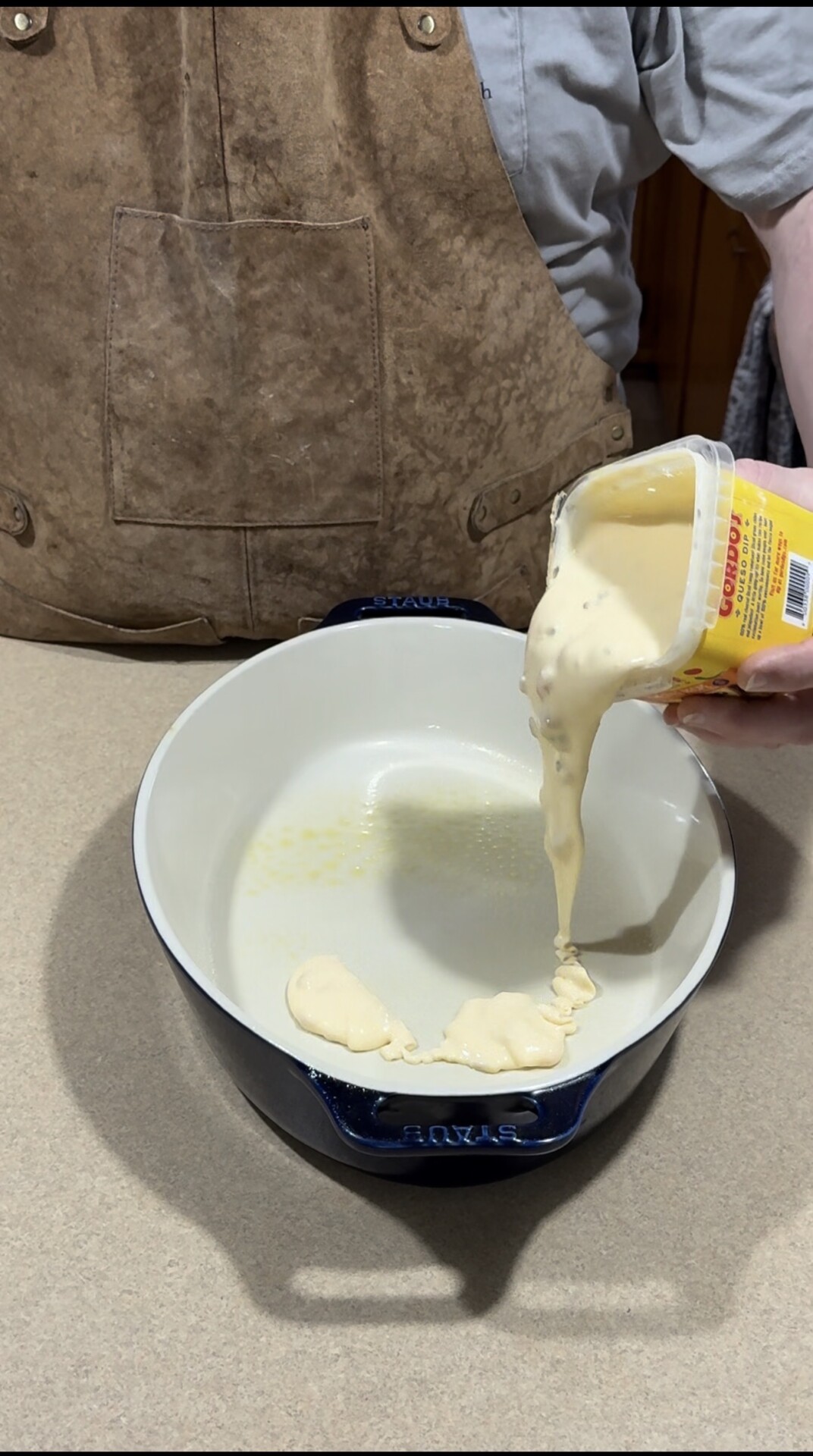 A person in a brown apron is pouring thick, creamy soup or sauce from a can into a white, oval baking dish on a kitchen counter.