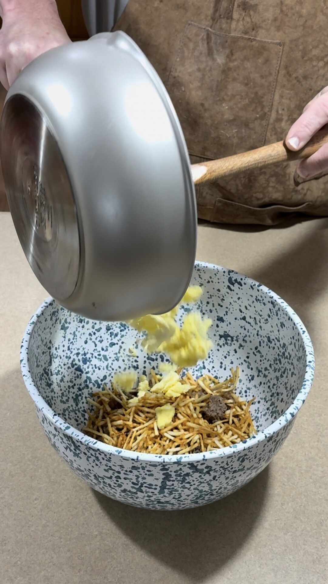 A person pours scrambled eggs from a silver pan into a speckled mixing bowl filled with crispy noodles and ground beef.