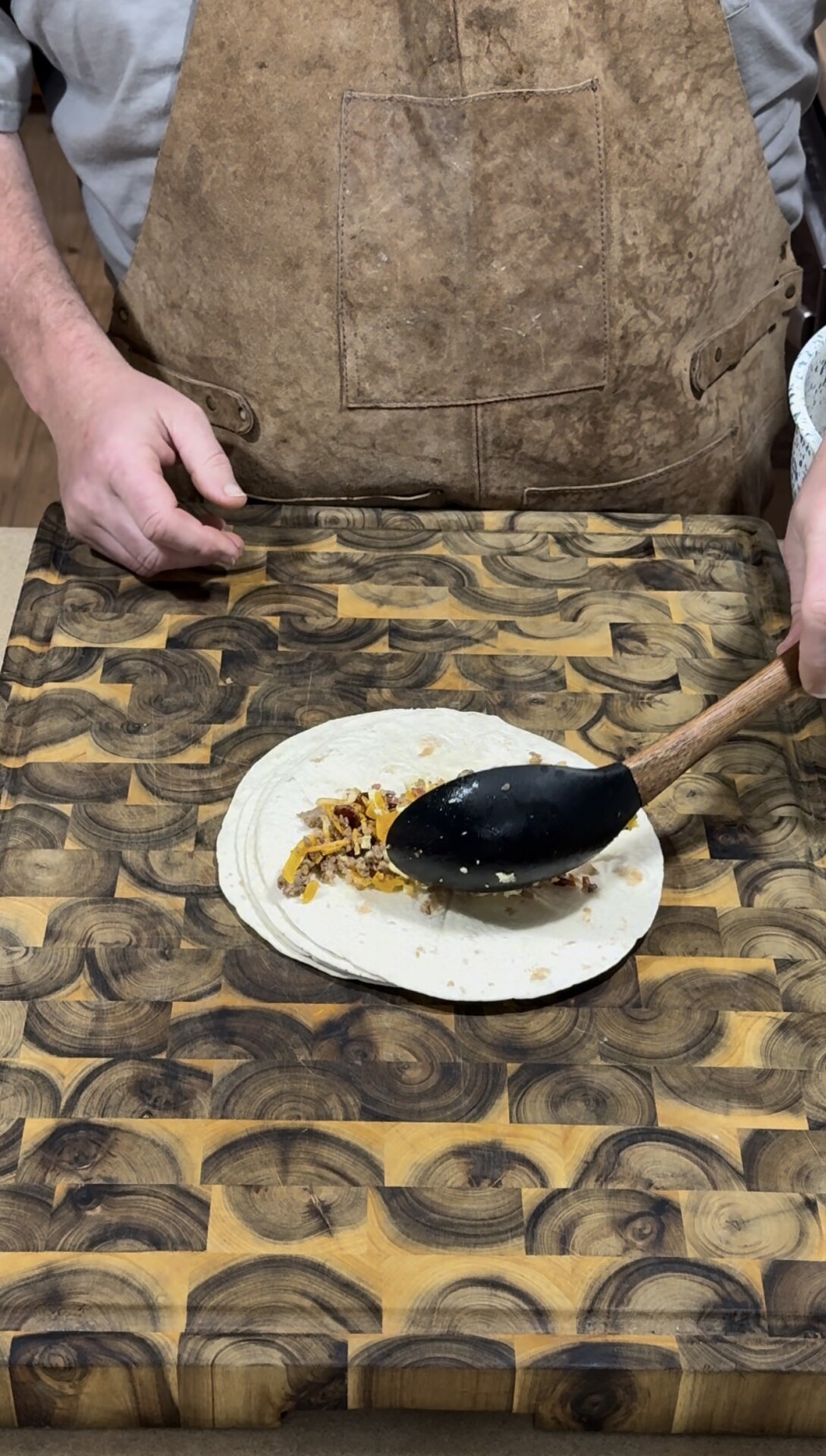 A person in a brown apron spoons a filling onto a tortilla wrap placed on a patterned wooden cutting board.