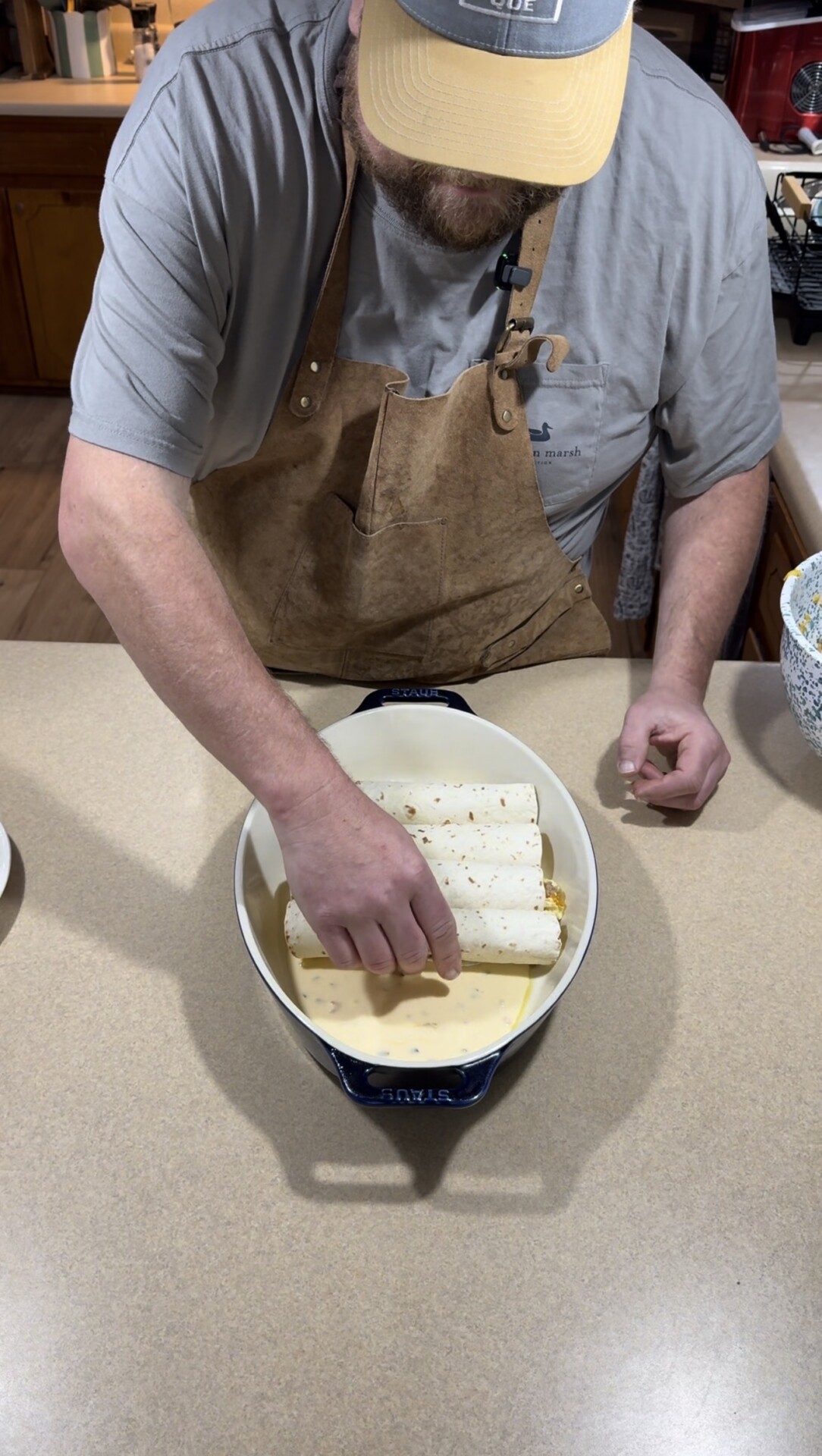 A person wearing a brown apron and gray shirt is placing rolled tortillas into a baking dish filled with sauce on a kitchen counter.