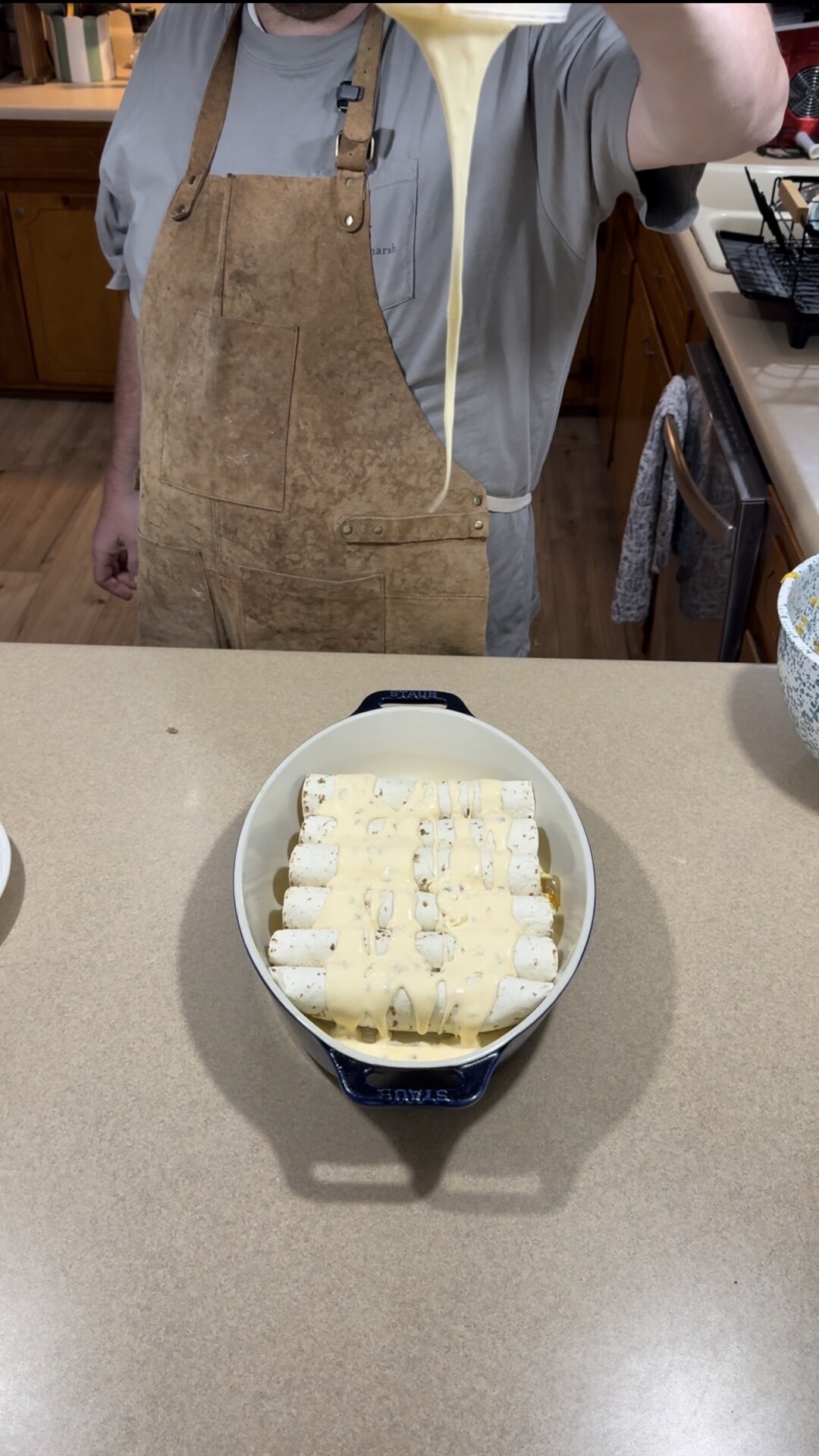 A person in a brown apron pours creamy sauce over a dish of rolled tortillas arranged in a baking dish on a kitchen counter.
