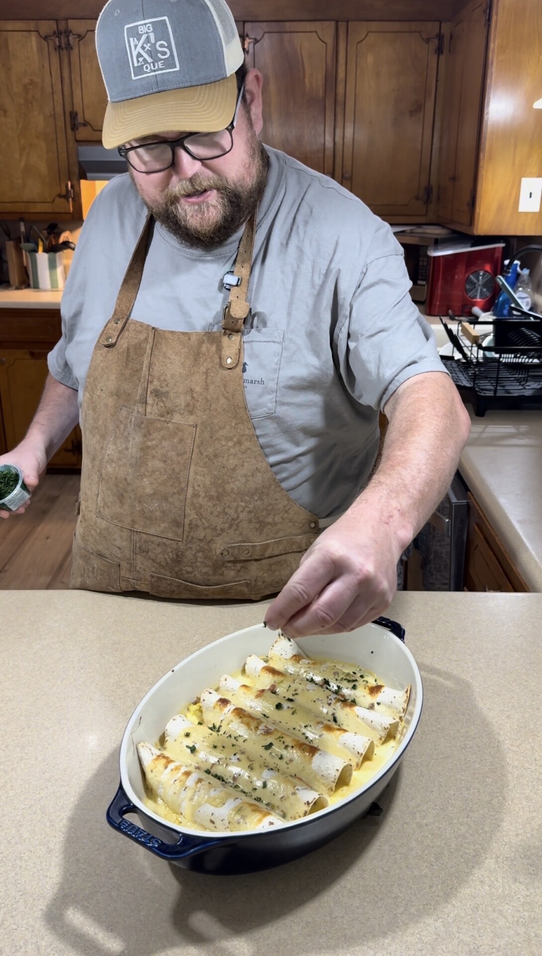 A man wearing glasses, a cap, and a brown apron sprinkles herbs over a dish of enchiladas in a kitchen with wooden cabinets.