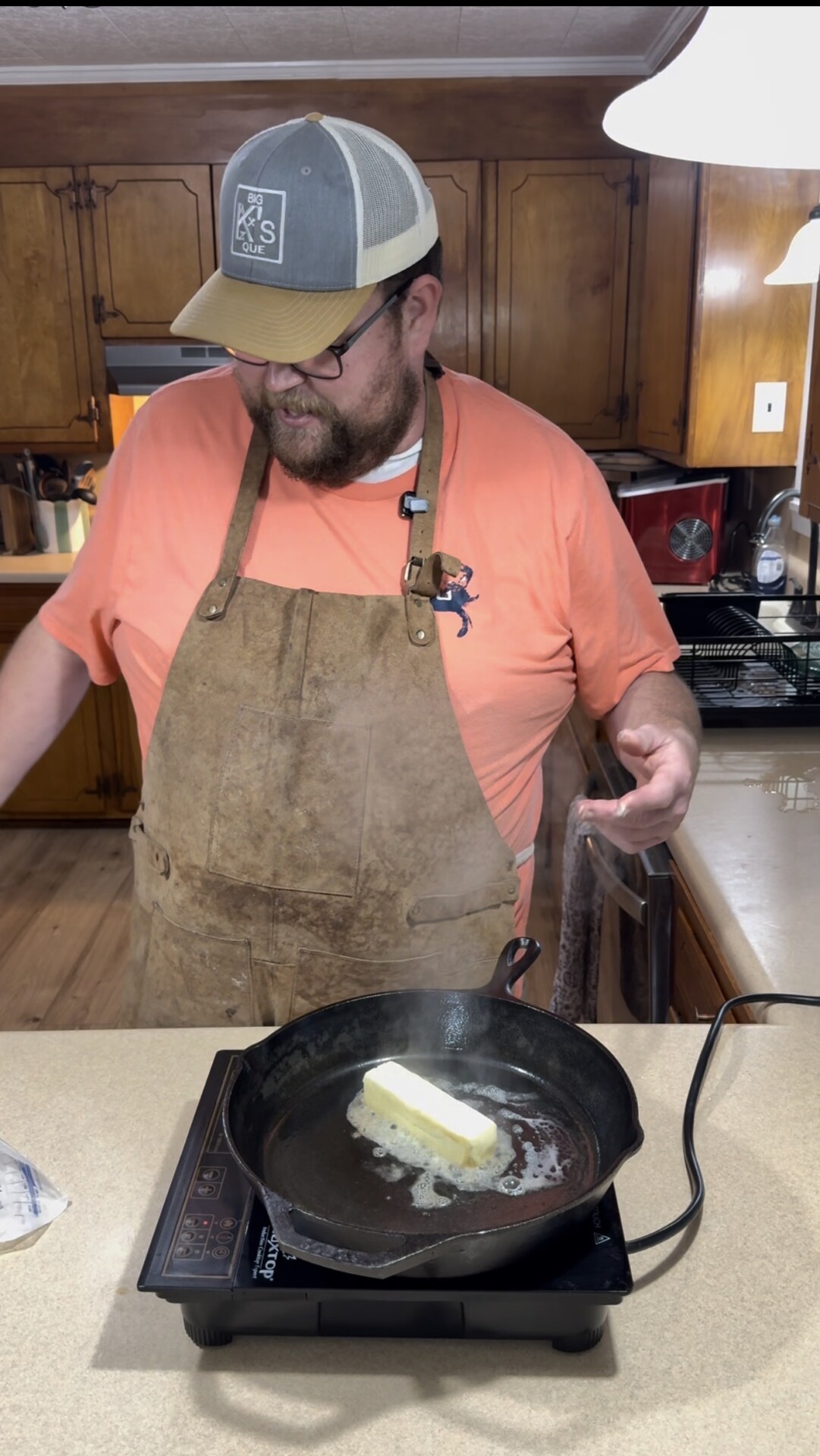 A man wearing a brown apron and a cap stands in a kitchen, looking down at a stick of butter melting in a black cast iron skillet on the stove. Wooden cabinets and kitchen appliances are in the background.