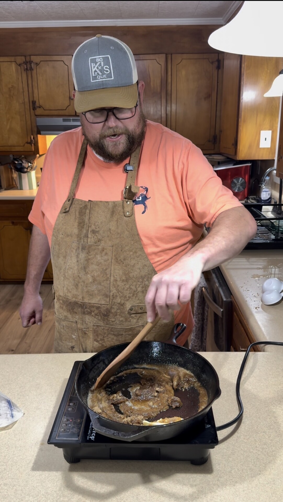A man wearing a cap, glasses, and a brown apron stirs a mixture in a cast iron skillet on a stovetop in a kitchen with wooden cabinets.