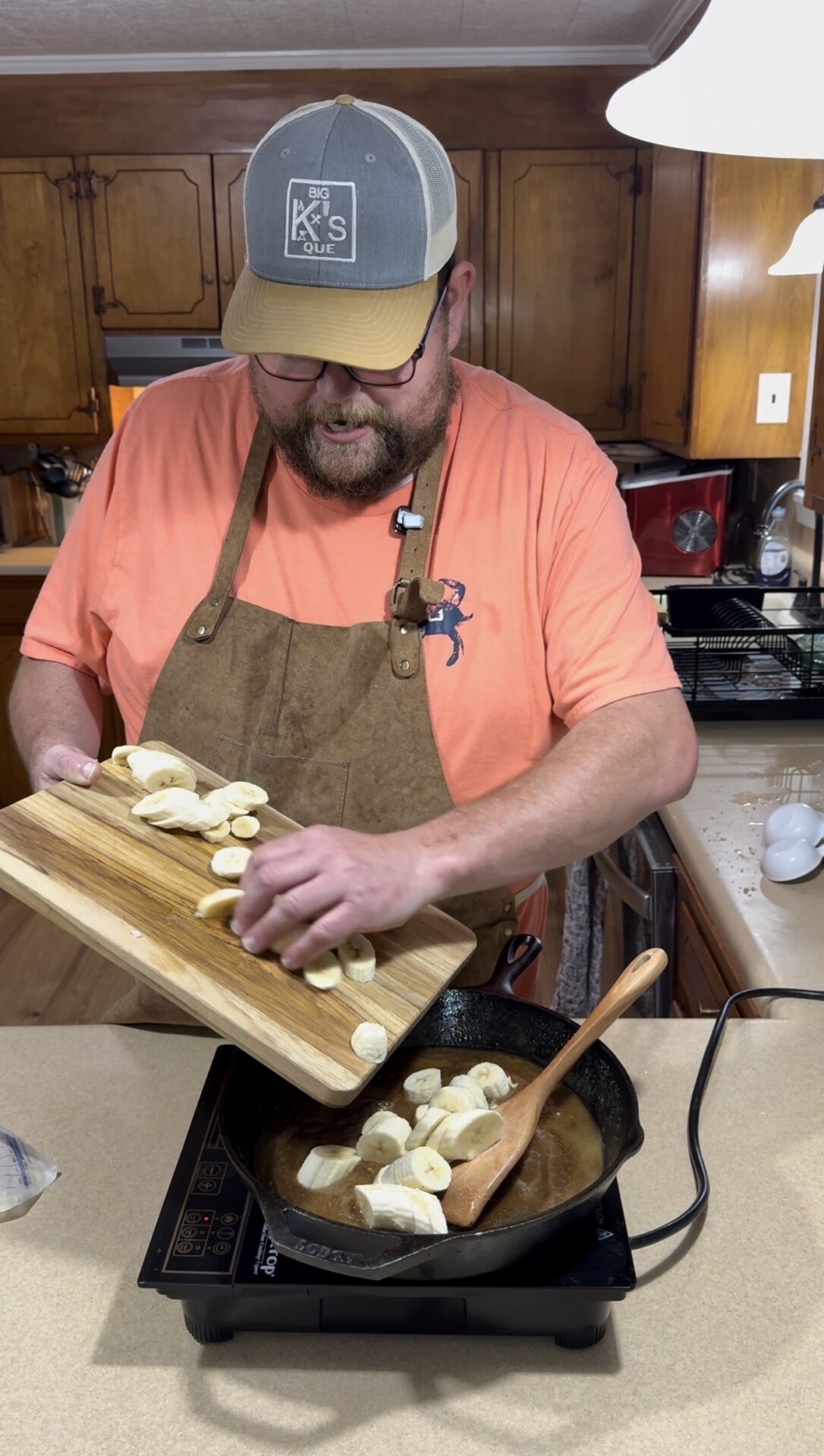 A man wearing a brown apron, orange shirt, and cap adds sliced bananas from a wooden cutting board into a skillet on the stove in a kitchen. A wooden spoon rests in the pan with simmering liquid.