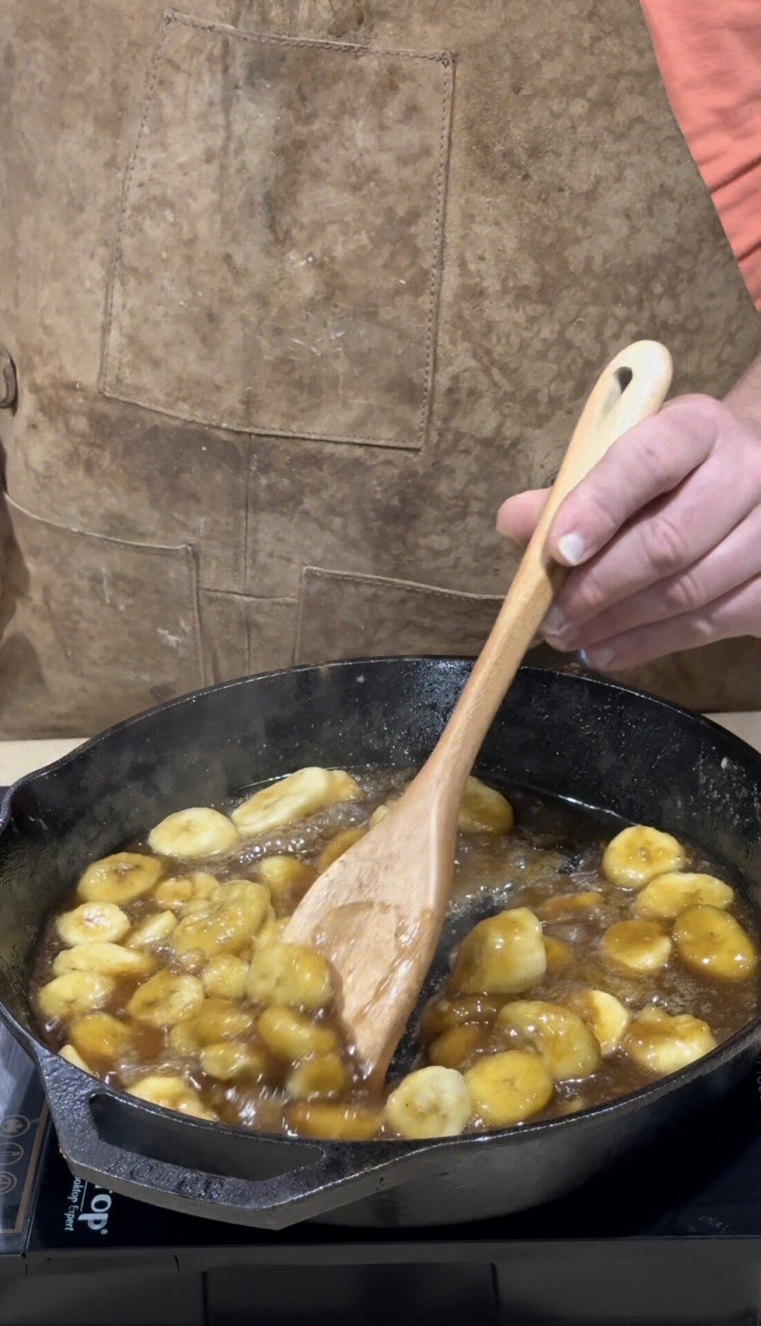 A person in a brown apron stirs sliced bananas in a bubbling caramel sauce with a wooden spoon in a black skillet.