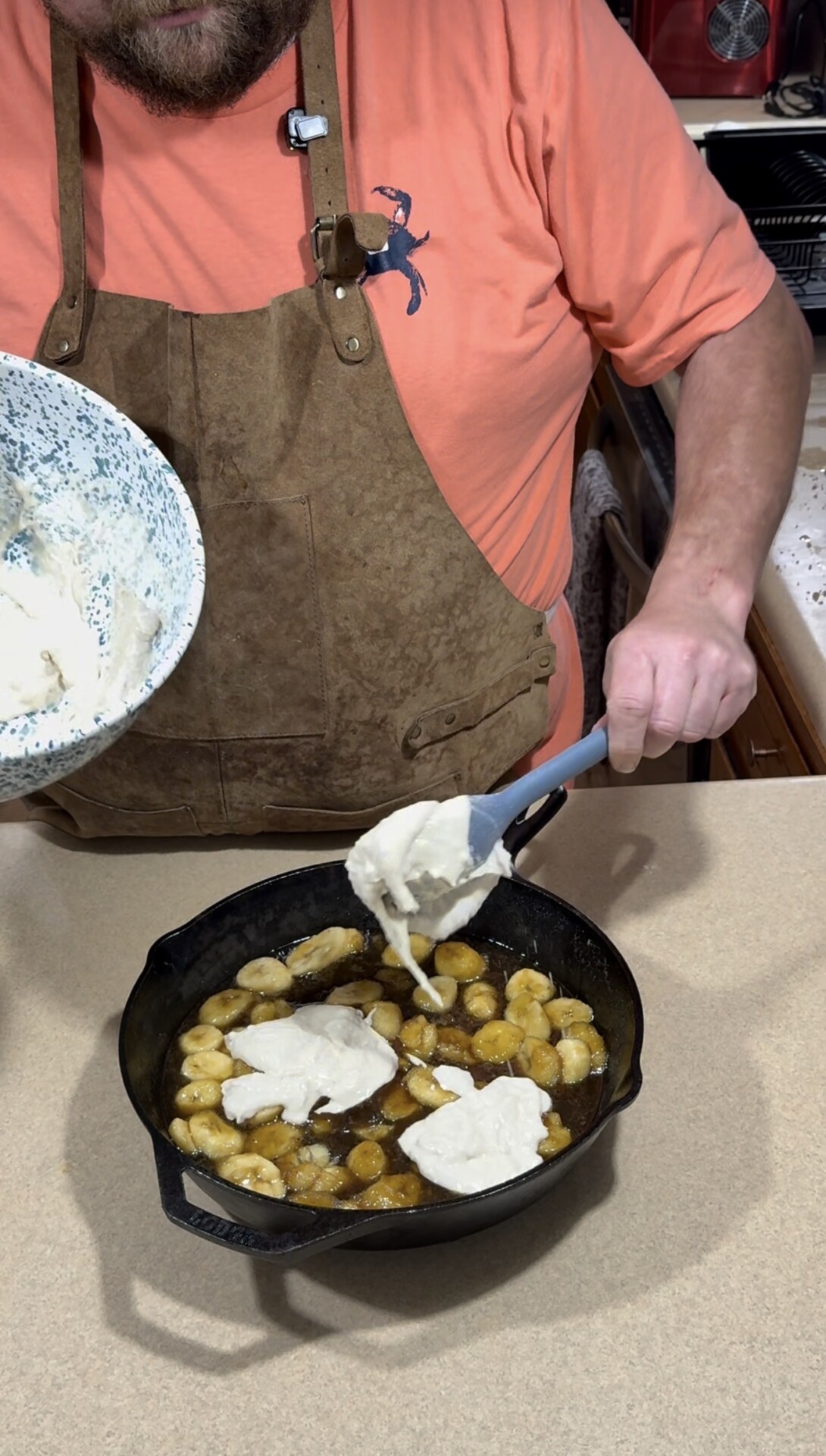 A person wearing a brown apron and orange shirt spoons biscuit dough from a bowl onto caramelized bananas in a cast iron skillet on a kitchen counter.